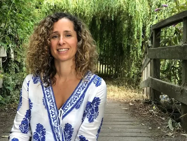 Cara Jordan, Birth and Postnatal Doula in London and South East England, sits next to the Thames in London, smiling and wearing a white blouse with a blue print. There is green foliage and a wooden bridge behind her.