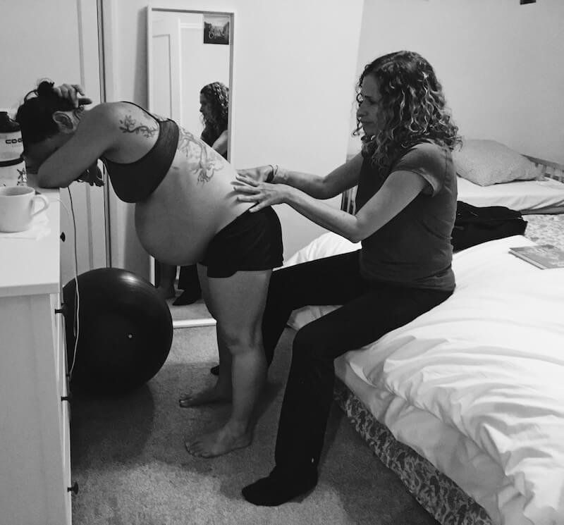 A woman in labour leans against a chest of drawers, Cara Jordan, a doula in London, massages the woman's back, a birth comfort technique.