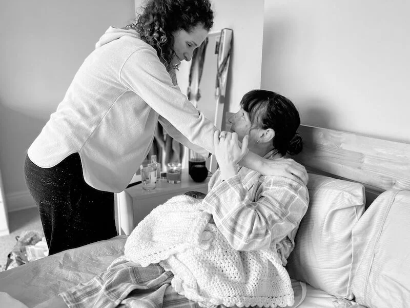 A doula smiles and exchanges warm words with her client in Chiswick. The client has just birthed her baby, the newborn baby rests in her mother's arms.