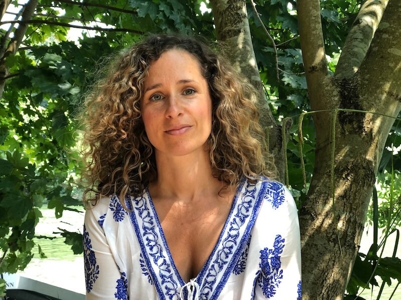 Cara Jordan, Birth Doula, Postnatal Doula and Night Nanny, sitting in front of some green trees in London. Cara has curly brown hair and wears a white blouse with a blue print.