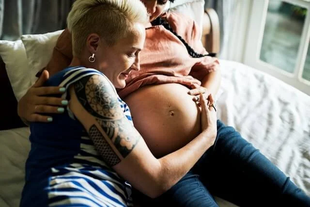 A pregnant woman is reclining on the bed, her female partner is cuddling her belly in Reading.