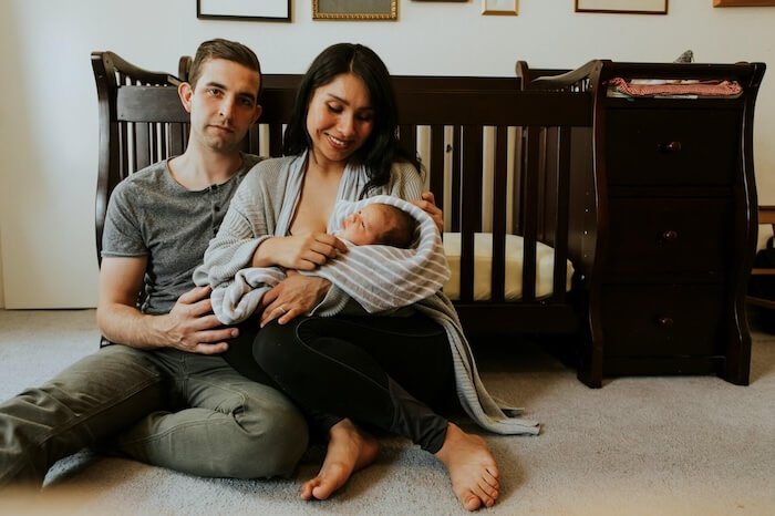 A couple sit on the floor cuddling their newborn baby in W14, London, the baby’s cot is behind them.
