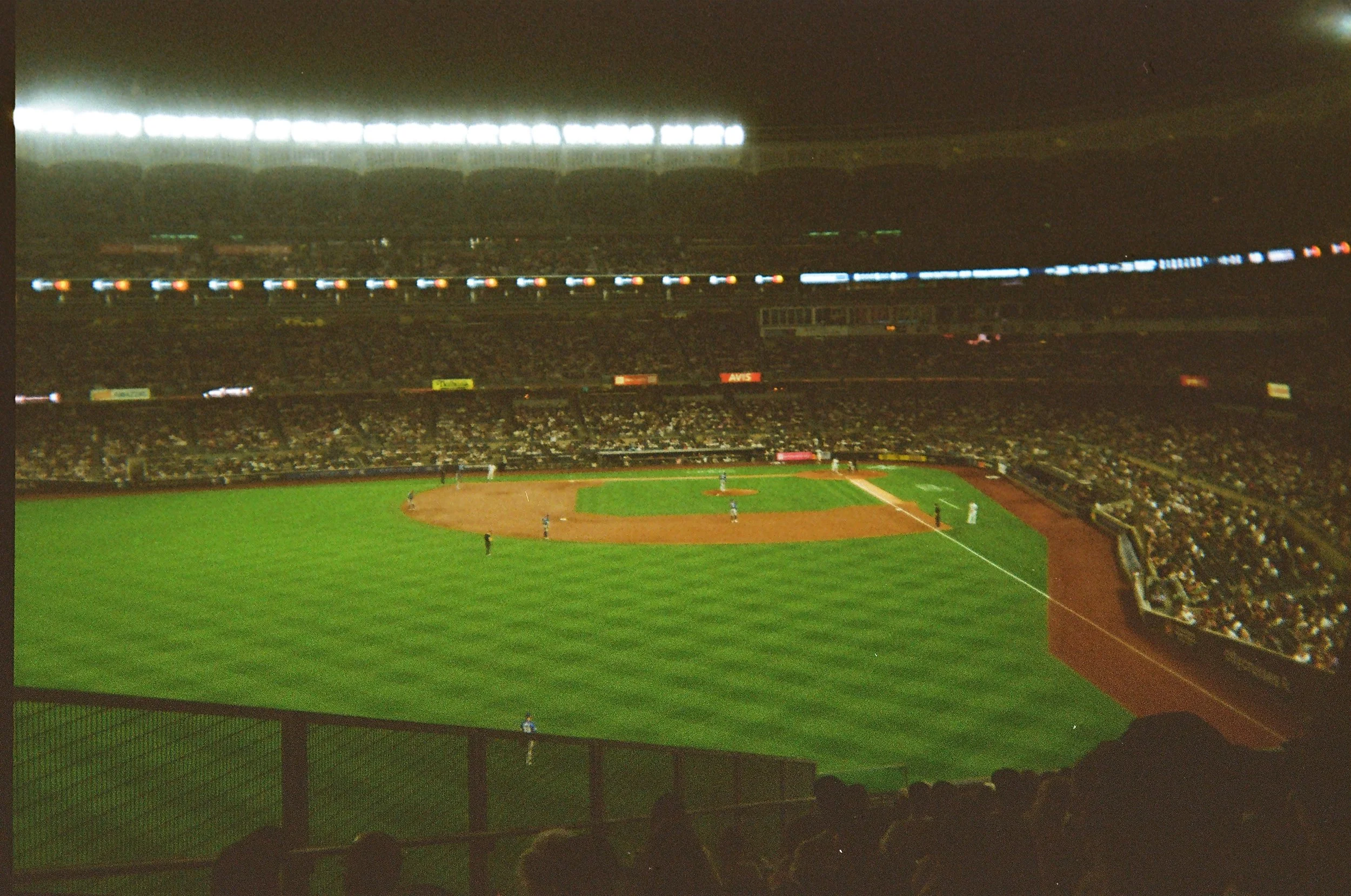 Nighttime baseball game at a large stadium with players on the field and spectators in the stands, illuminated by stadium lights.