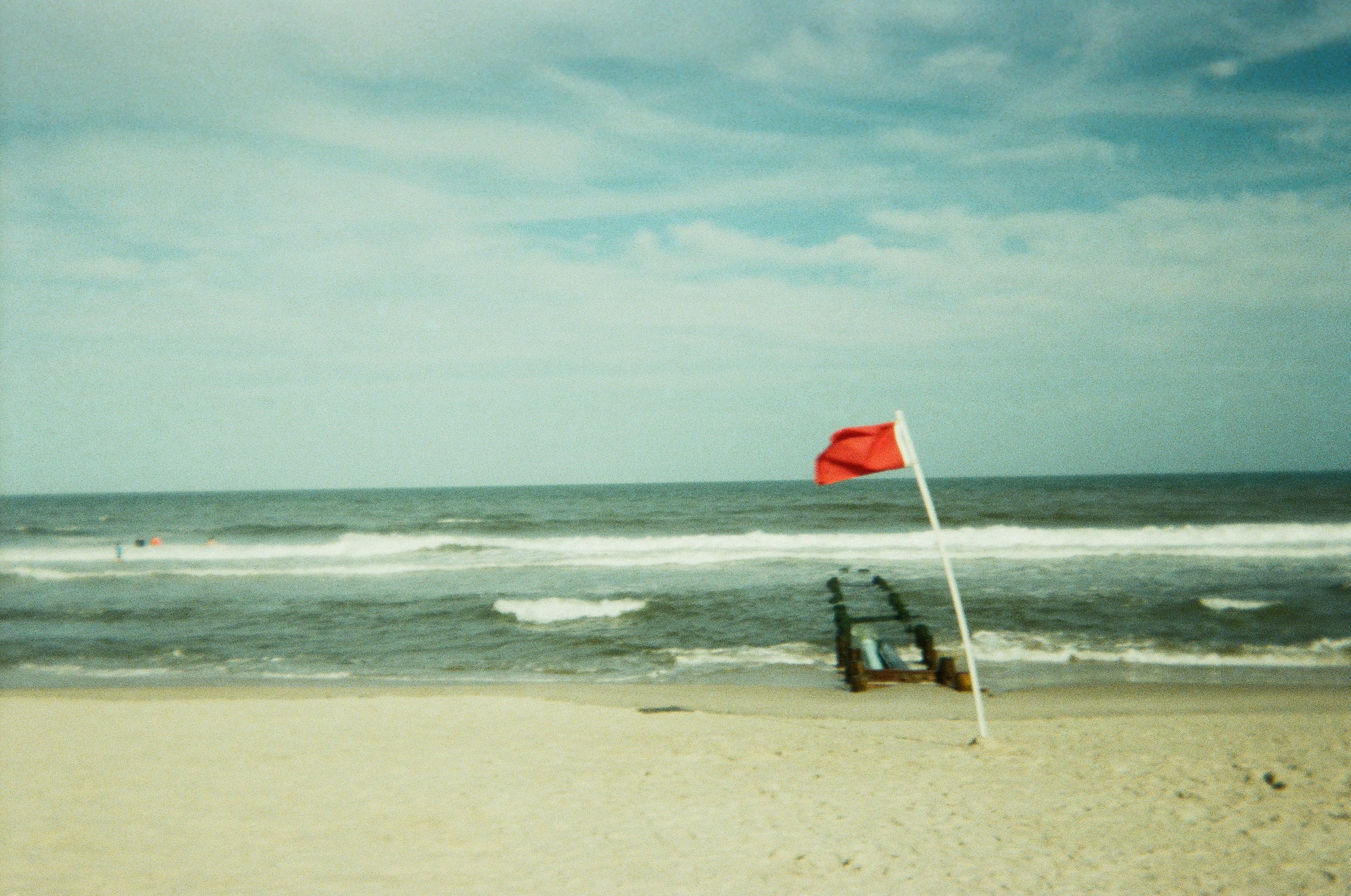 Empty beach with a red flag and a wooden lifeguard chair on the sandy shore, with ocean waves and a cloudy sky in the background.