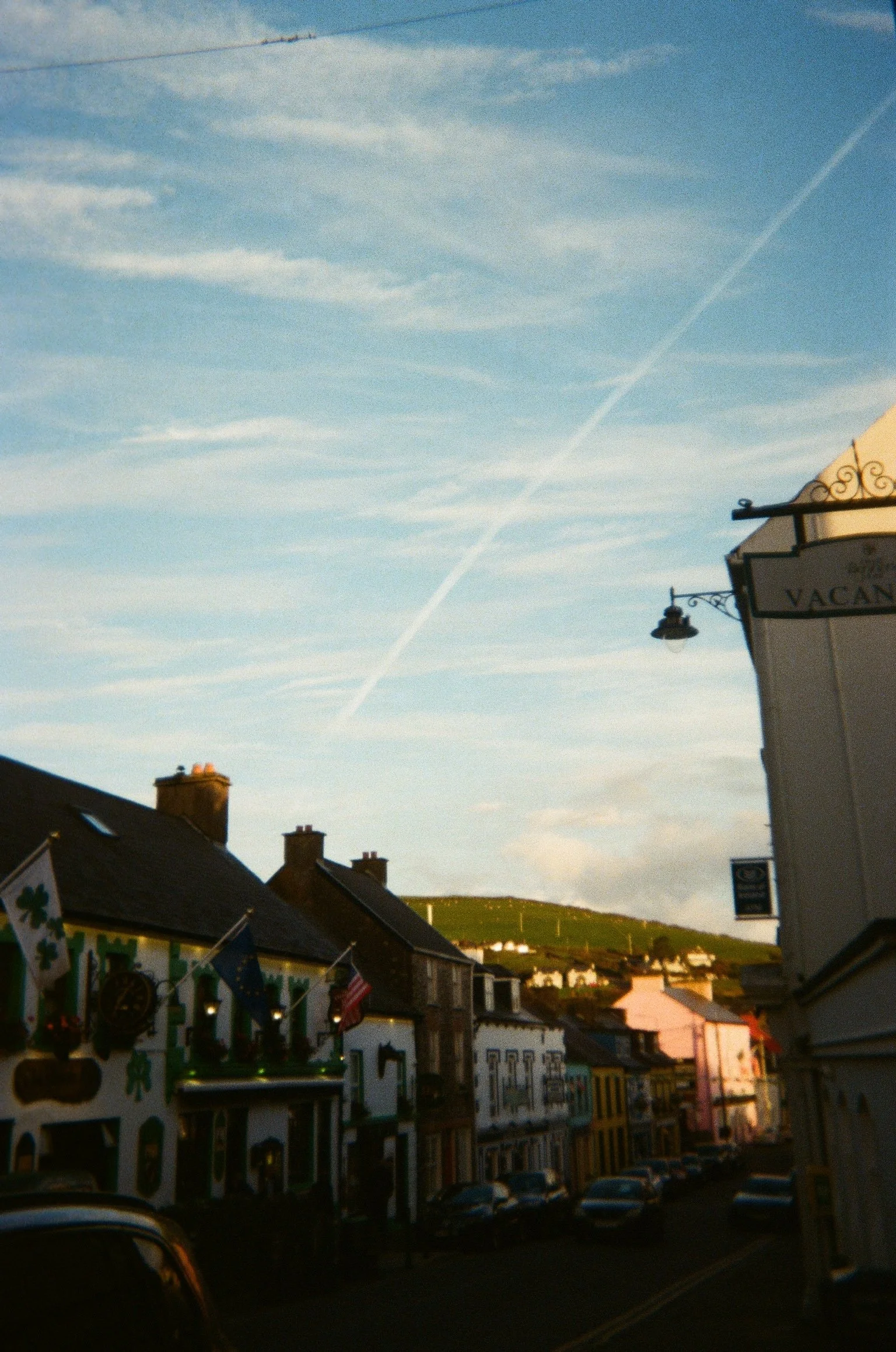 A street view of a small town featuring colorful buildings, some decorated with flags, parked cars, a lamp post, and a sky with wispy clouds and a contrail.