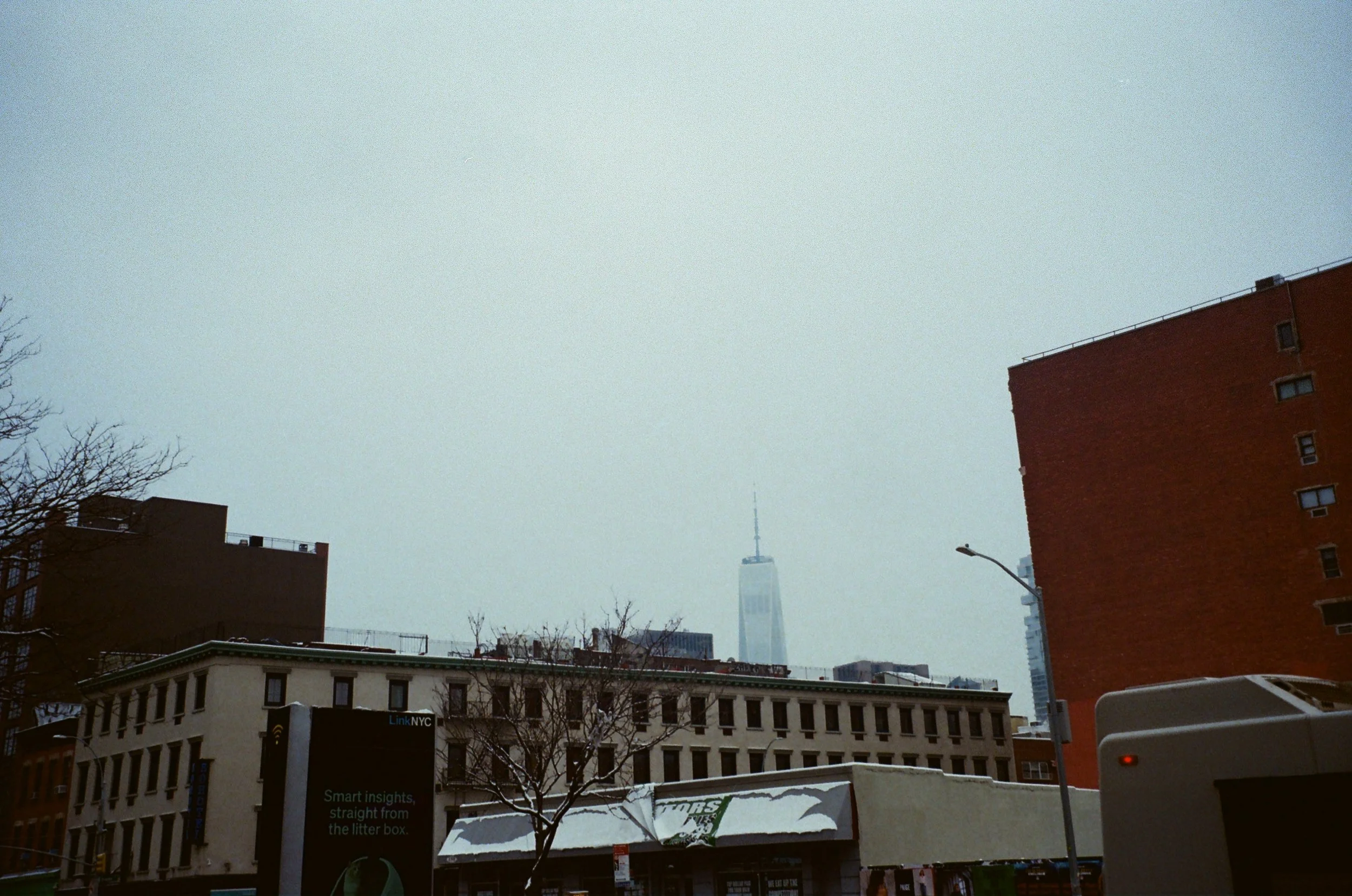 A cityscape featuring a view of a tall building, possibly the One World Trade Center, in the distance, with shorter buildings in the foreground and a street with bare trees and a bus in the lower part of the image.