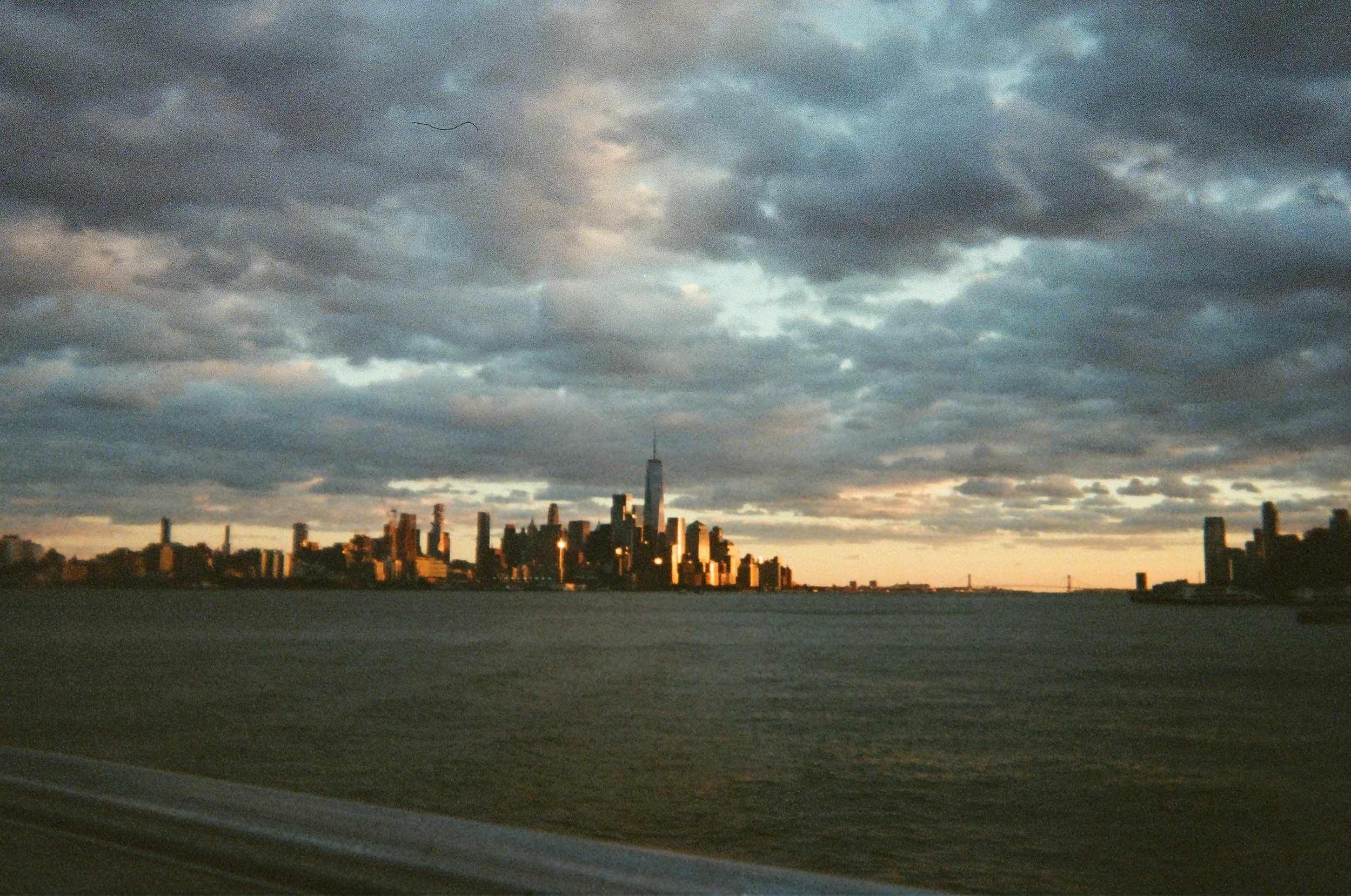 View of the Manhattan skyline at sunset, with tall buildings including One World Trade Center, viewed across a body of water with cloudy sky.