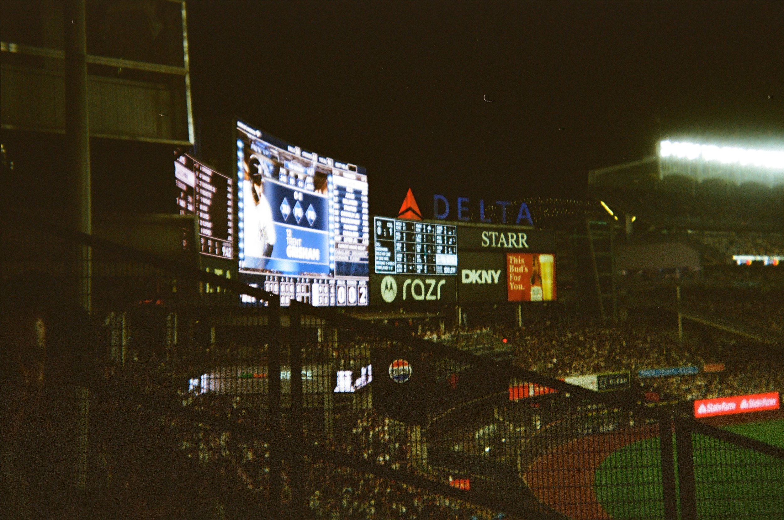 Nighttime view of a baseball stadium with illuminated scoreboard displaying player information, advertisements, and the stadium logo, and a crowd in the stands.