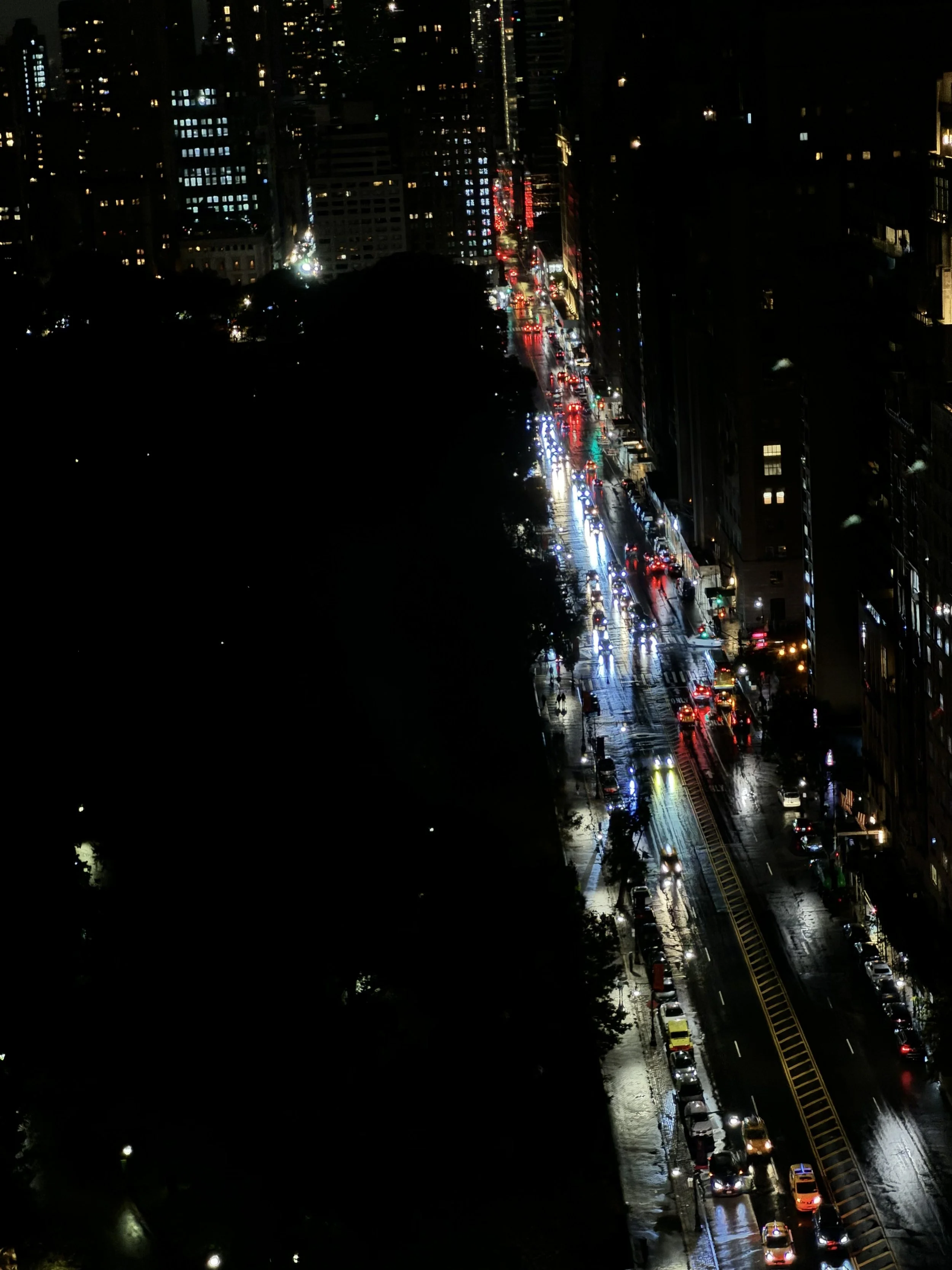 Nighttime city street scene with wet roads, parked cars, vehicles moving, illuminated buildings, and reflections on the wet pavement.