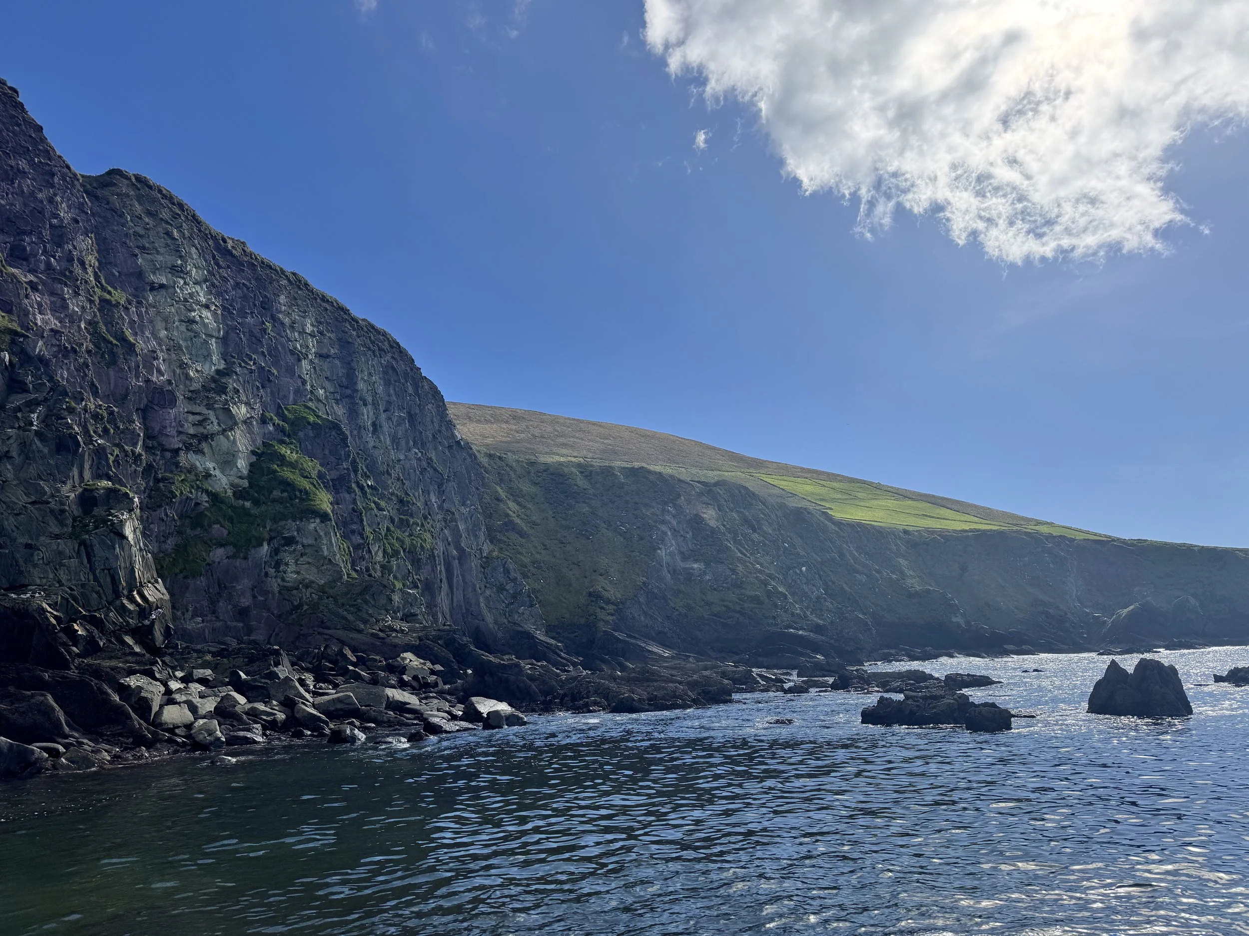 Irish landscape with rocky cliffs on the left, grassy slopes on the right, and a body of water in the foreground under a partly cloudy blue sky.