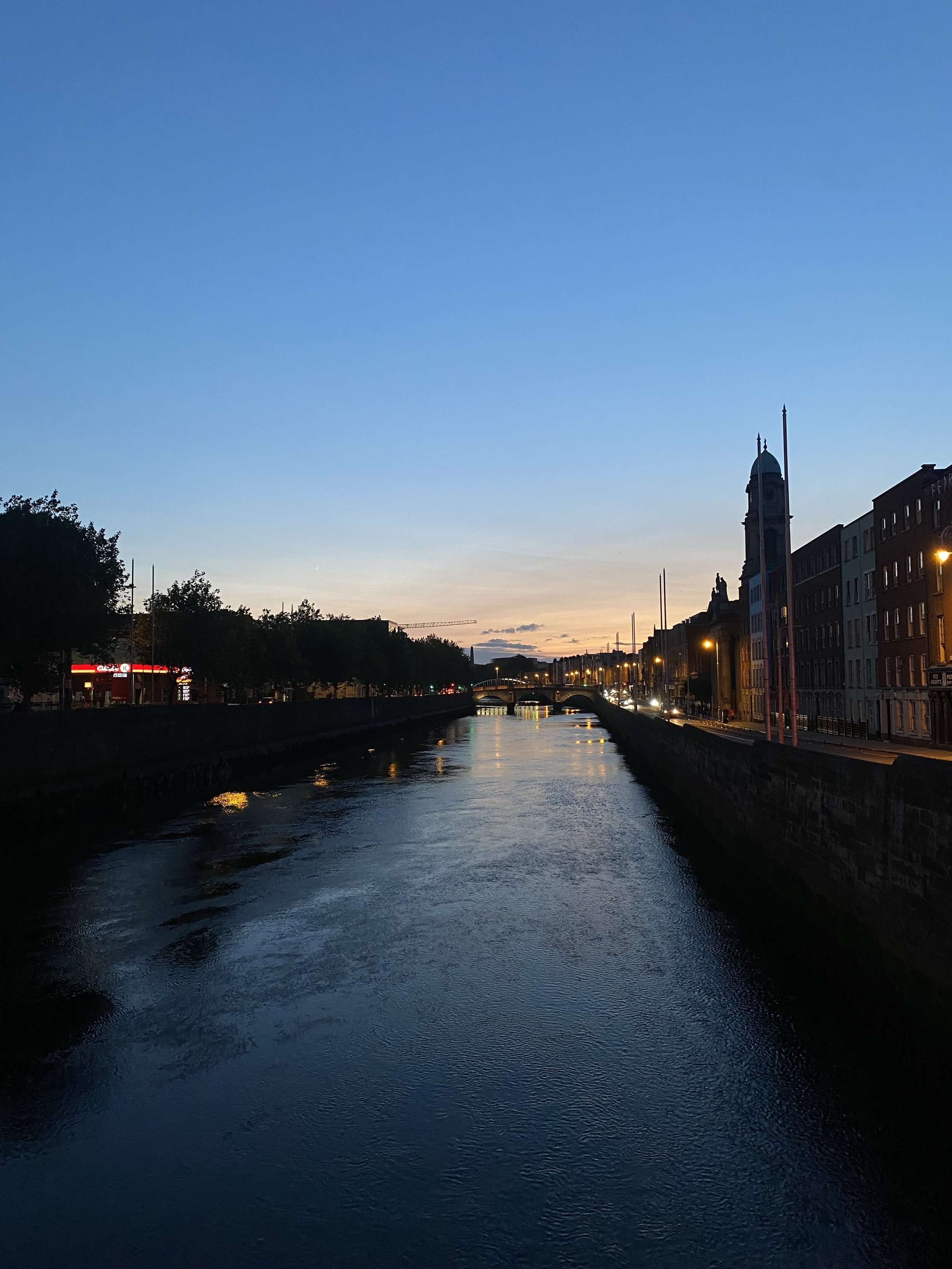 Dublin city canal at dusk with buildings and a church tower on the right, streetlights reflecting on the water, and a bridge in the distance against a fading sunset sky.