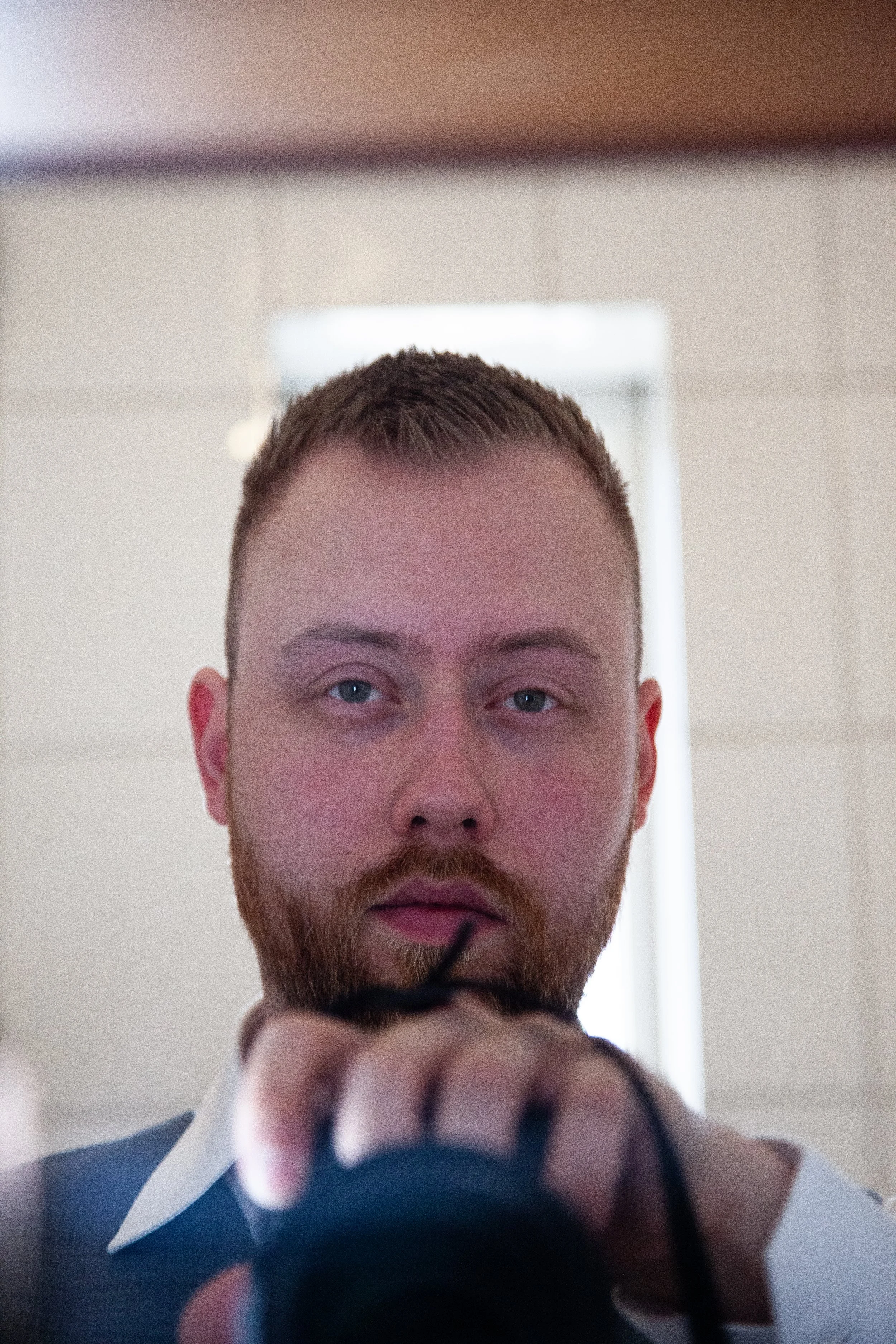A man with short brown hair and a beard, wearing a suit, is taking a selfie in front of a mirror, holding a camera.