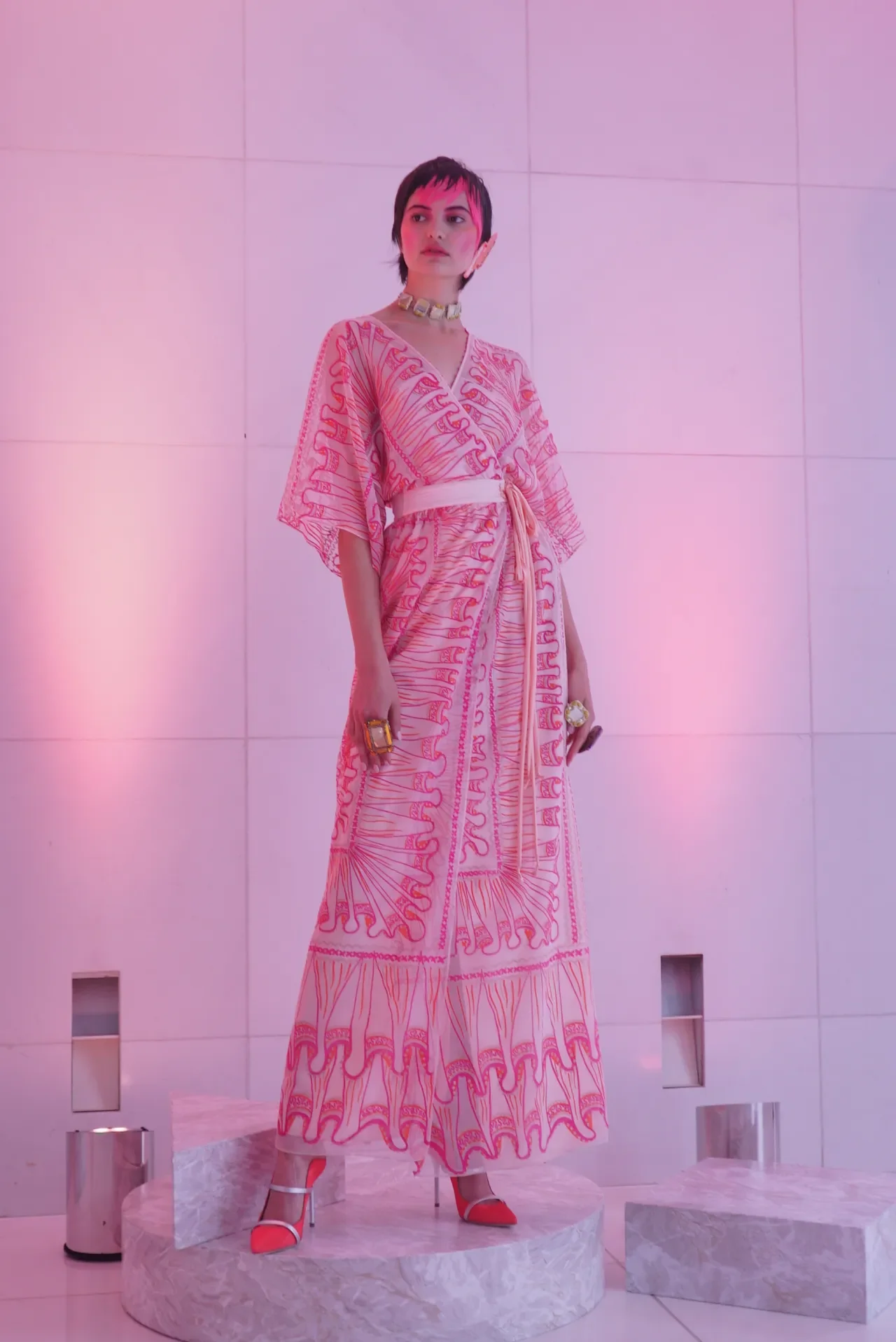 A woman in a pink and white patterned dress standing on a marble platform in front of a pink-lit tiled wall, wearing jewelry and heels.