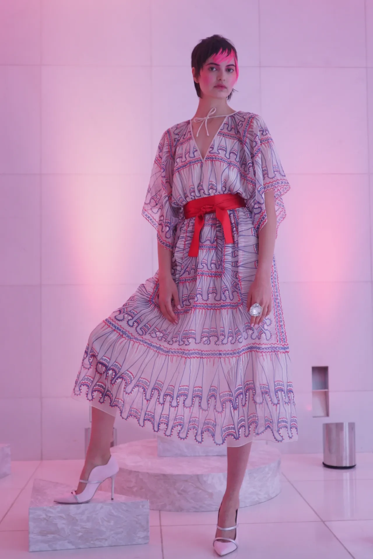 A woman with short dark hair in a patterned dress with a red waist sash and white high heels, standing against a pink-lit wall.