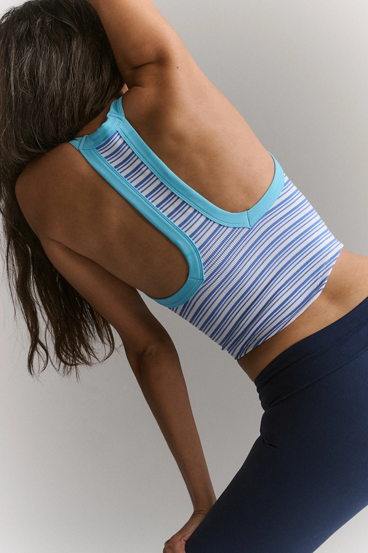 A woman wearing a striped tank top and dark pants, stretching with her arm over her head.