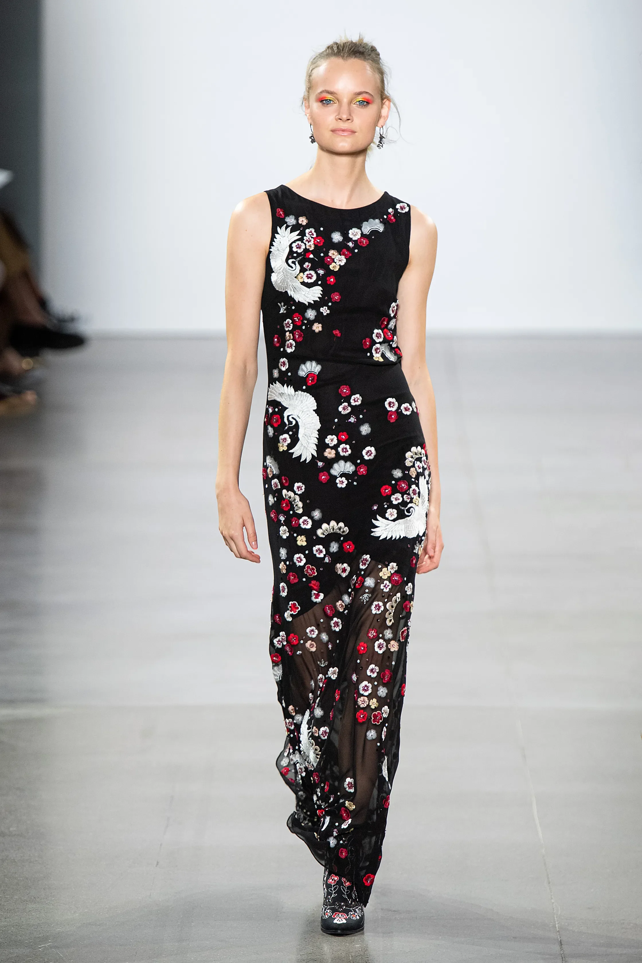 A female model walking down a runway wearing a black sheer dress with floral and bird embroidery.
