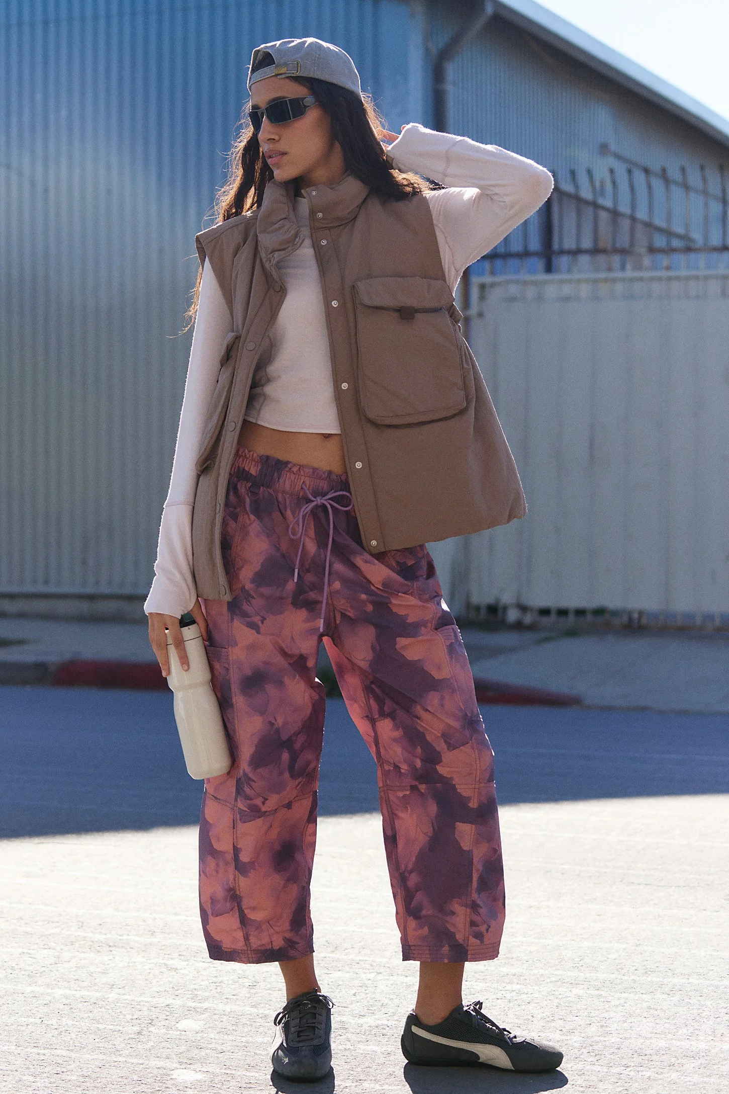 A woman with long dark hair wearing sunglasses, a backward gray baseball cap, a beige vest over a white long-sleeve shirt, camouflage pants, and black sneakers, standing outdoors in front of a corrugated metal building, holding a water bottle.