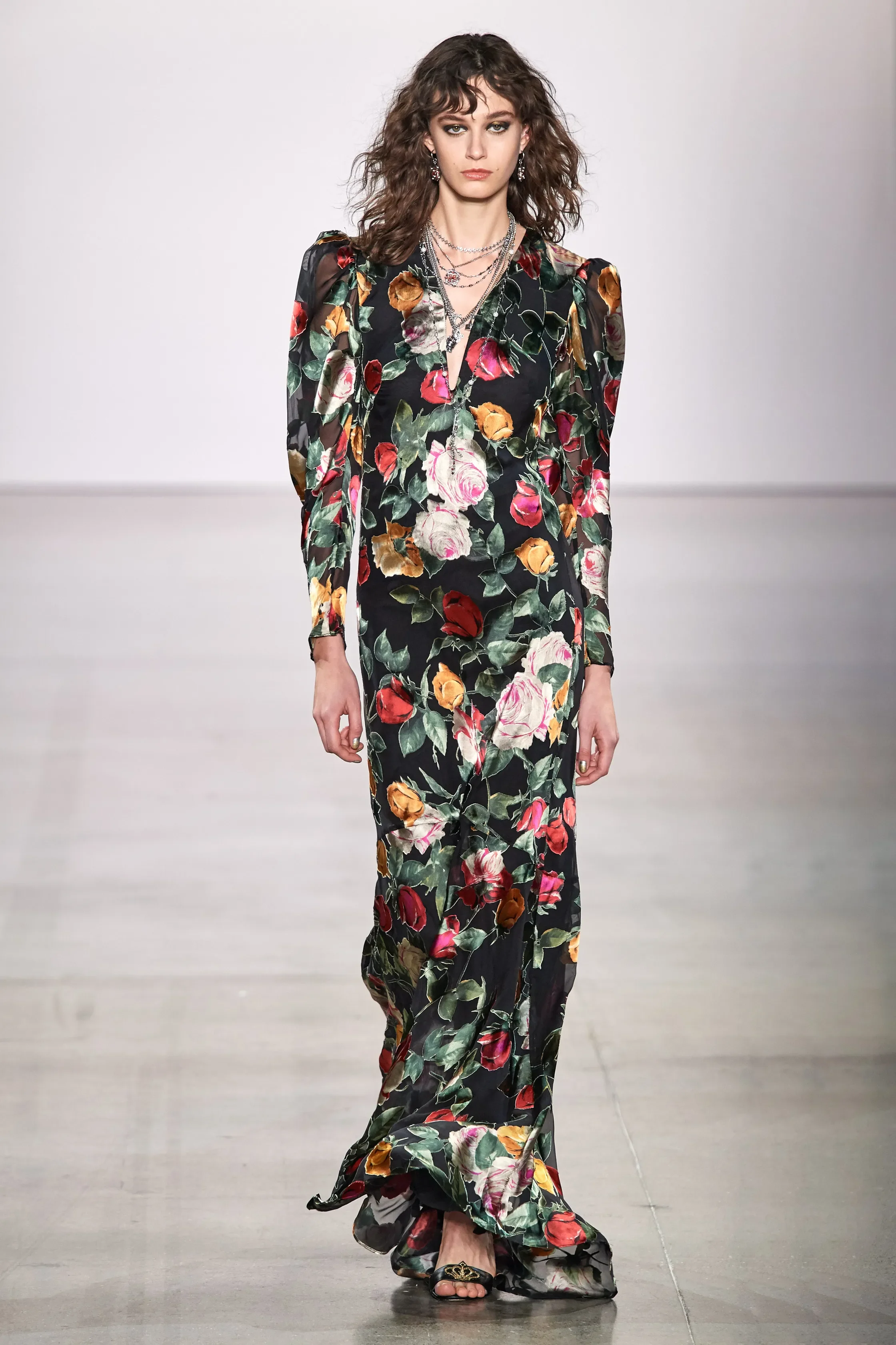 Female model walking down a runway in a long floral dress with black background and colorful flowers, accessorized with necklaces and earrings.