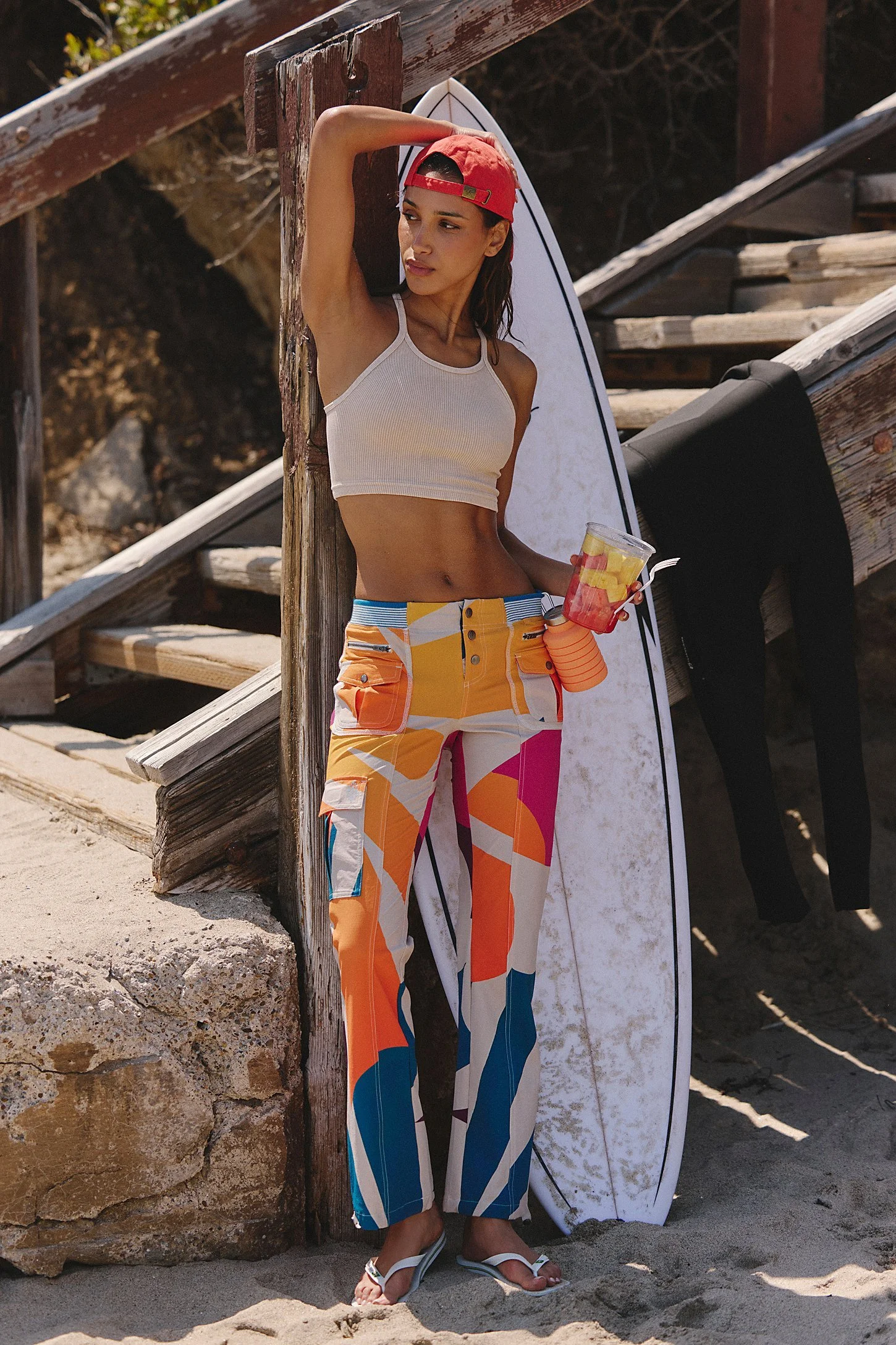 A woman standing on a sandy beach leaning against a wooden post. She is wearing a white crop top, colorful patterned pants, and a red backwards cap. She holds a drink with fruit in her right hand and is next to a surfboard.