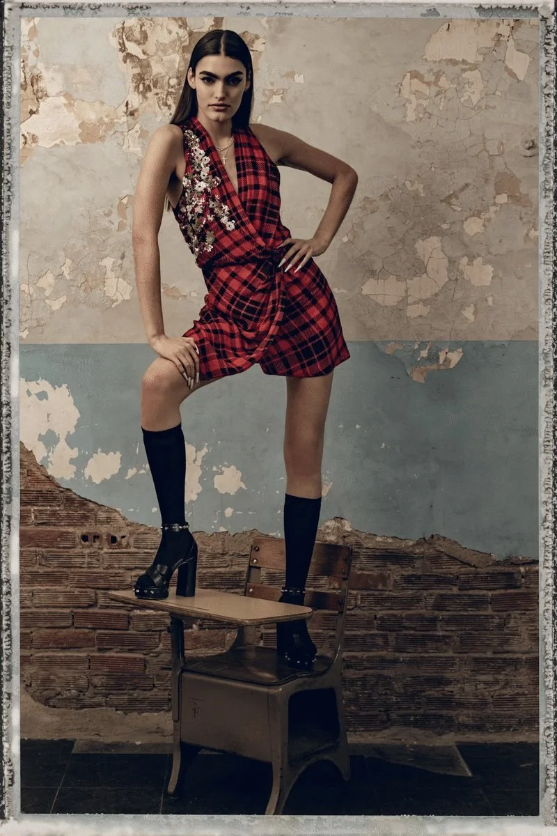 A woman standing with one foot on a chair and the other on a chair back, wearing a sleeveless red plaid dress with floral embellishments, black knee-high socks, and high-heeled shoes, in an industrial-style room with peeling paint on the walls.