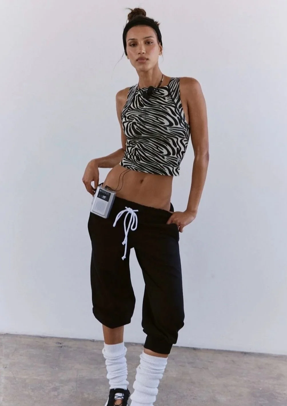 Woman in zebra print crop top, black sweatpants with white drawstring, white leg warmers, and black sneakers, standing against a plain white wall.