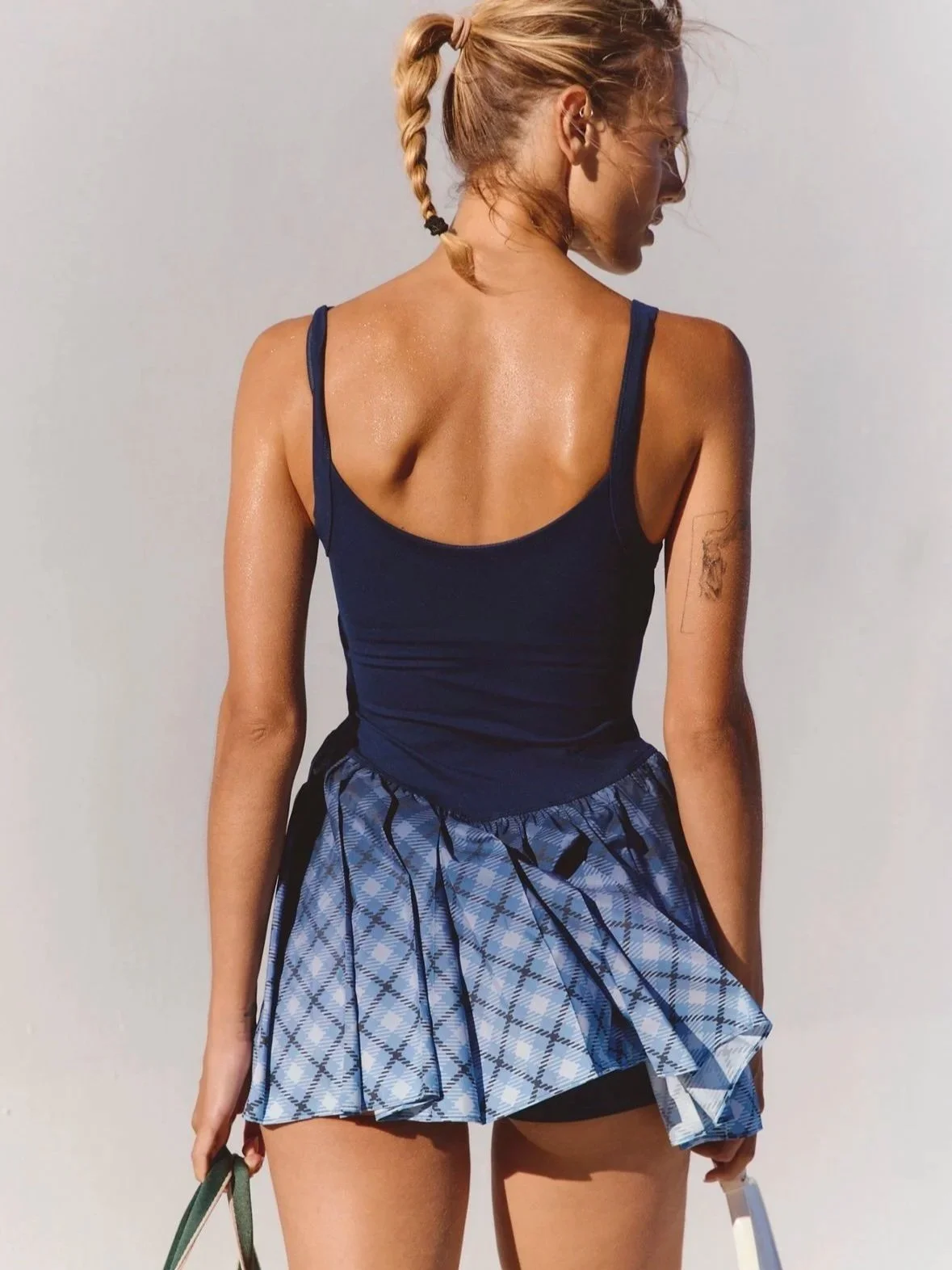 A woman with a braid, wearing a navy tank top and a blue plaid skirt, standing against a plain light-colored background.