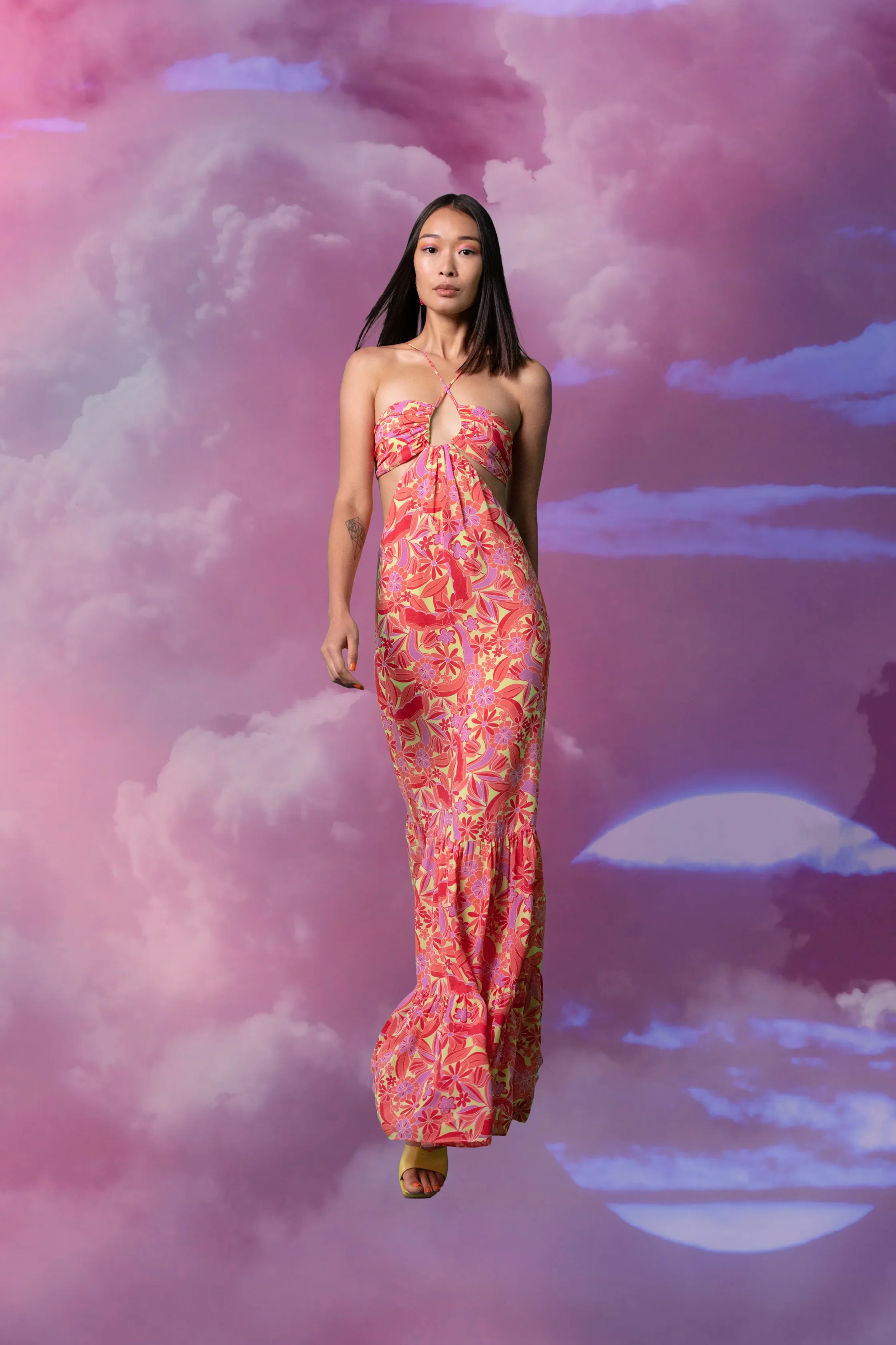 A woman in a colorful floral dress standing against a surreal sky background with clouds and a partial moon.