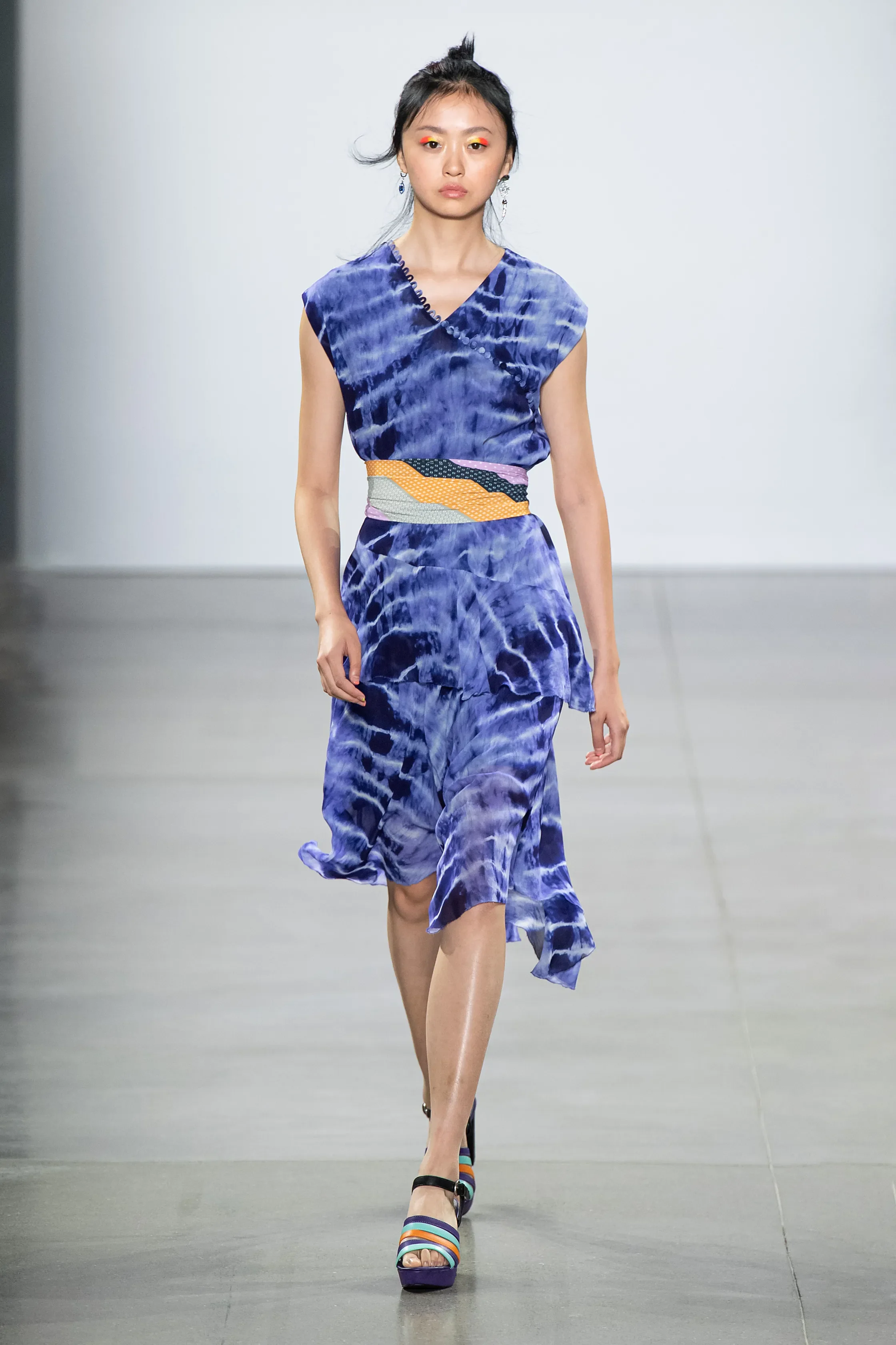 Model walking on runway wearing a blue tie-dye styled dress with ruffled hem, paired with colorful striped high heel sandals.