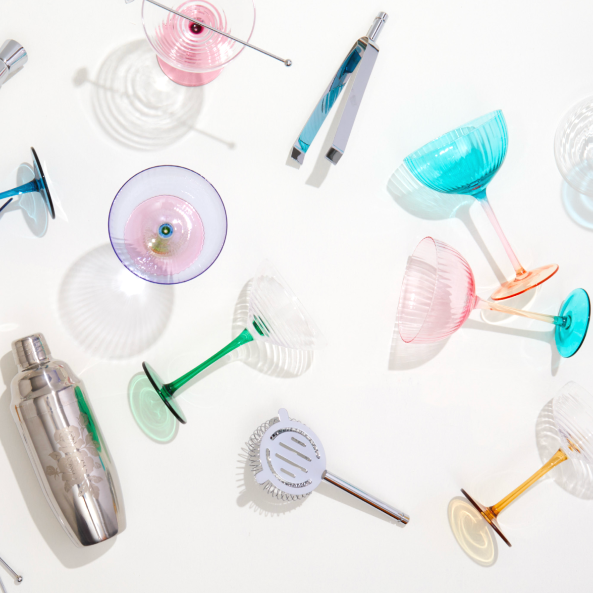 Colorful cocktail glasses and a cocktail shaker arranged on a white surface, with shadows cast by the glasses in a well-lit setting.