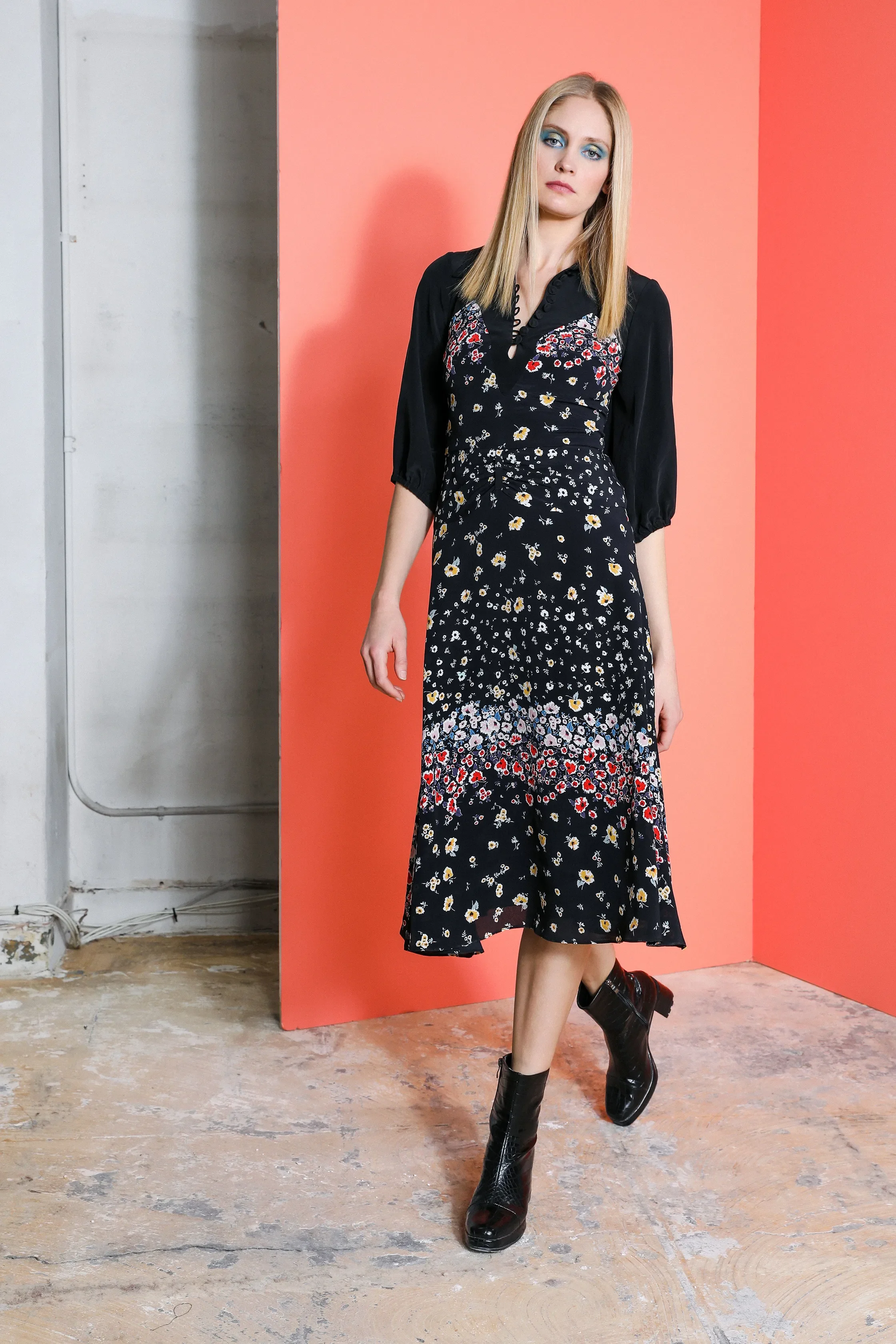 A young woman with blonde hair and blue eyeshadow wearing a black floral dress and black ankle boots, standing on a wooden floor with a coral-colored background.