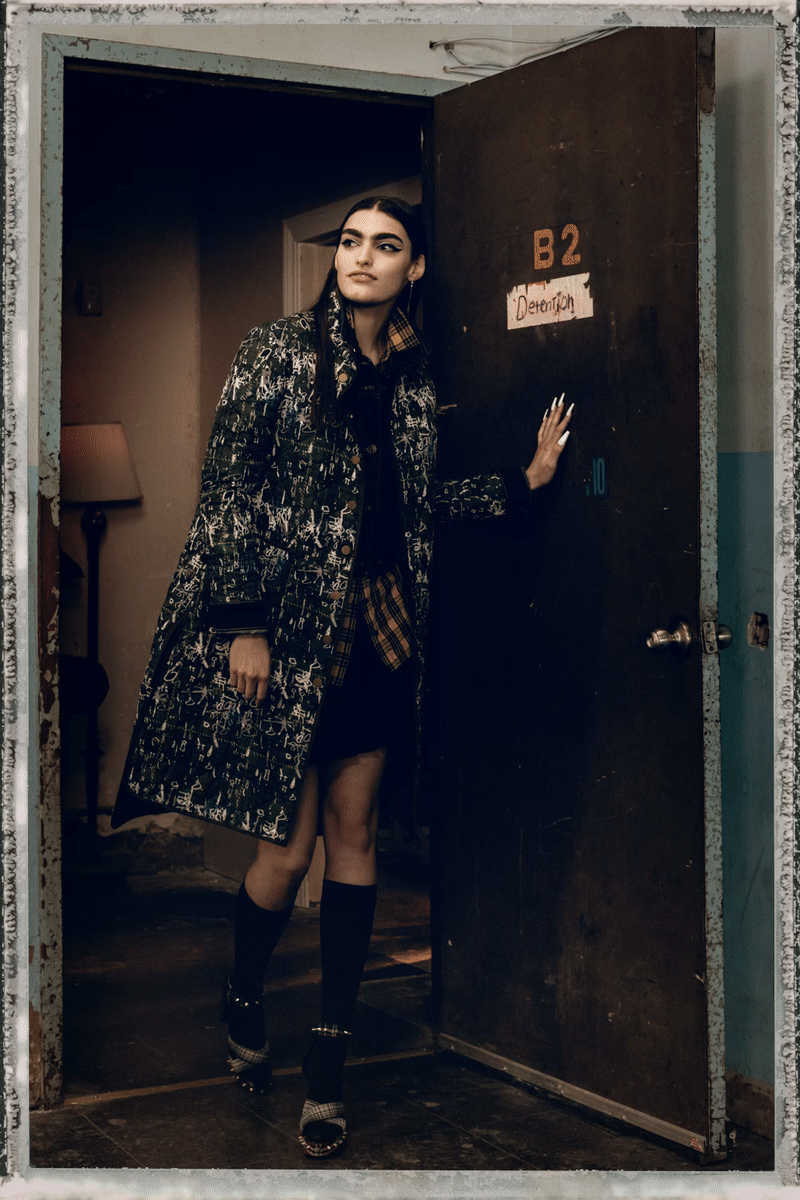 A woman with dark hair and a patterned coat standing in a doorway marked B2 Demolition, inside a dimly lit room.
