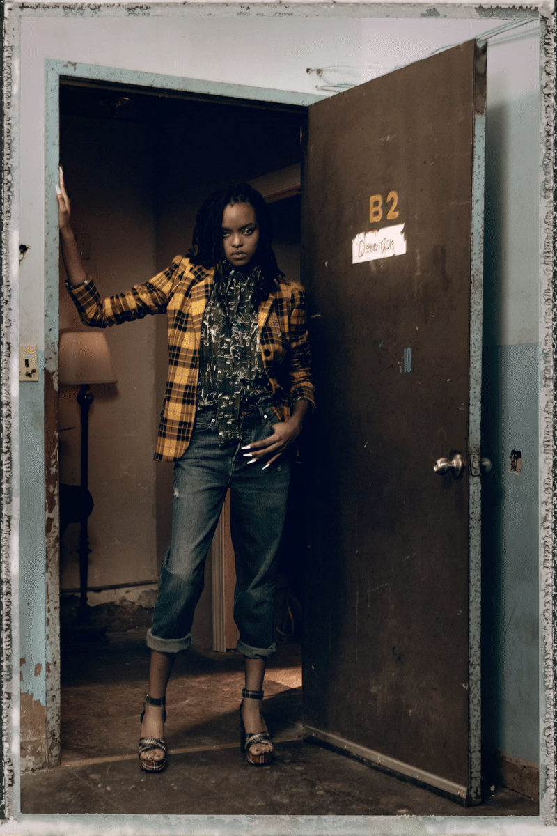 A woman standing in a doorway, wearing a yellow plaid blazer, a patterned blouse, jeans with rolled-up cuffs, and high heels, in a dimly lit room with an old lamp and worn decor.