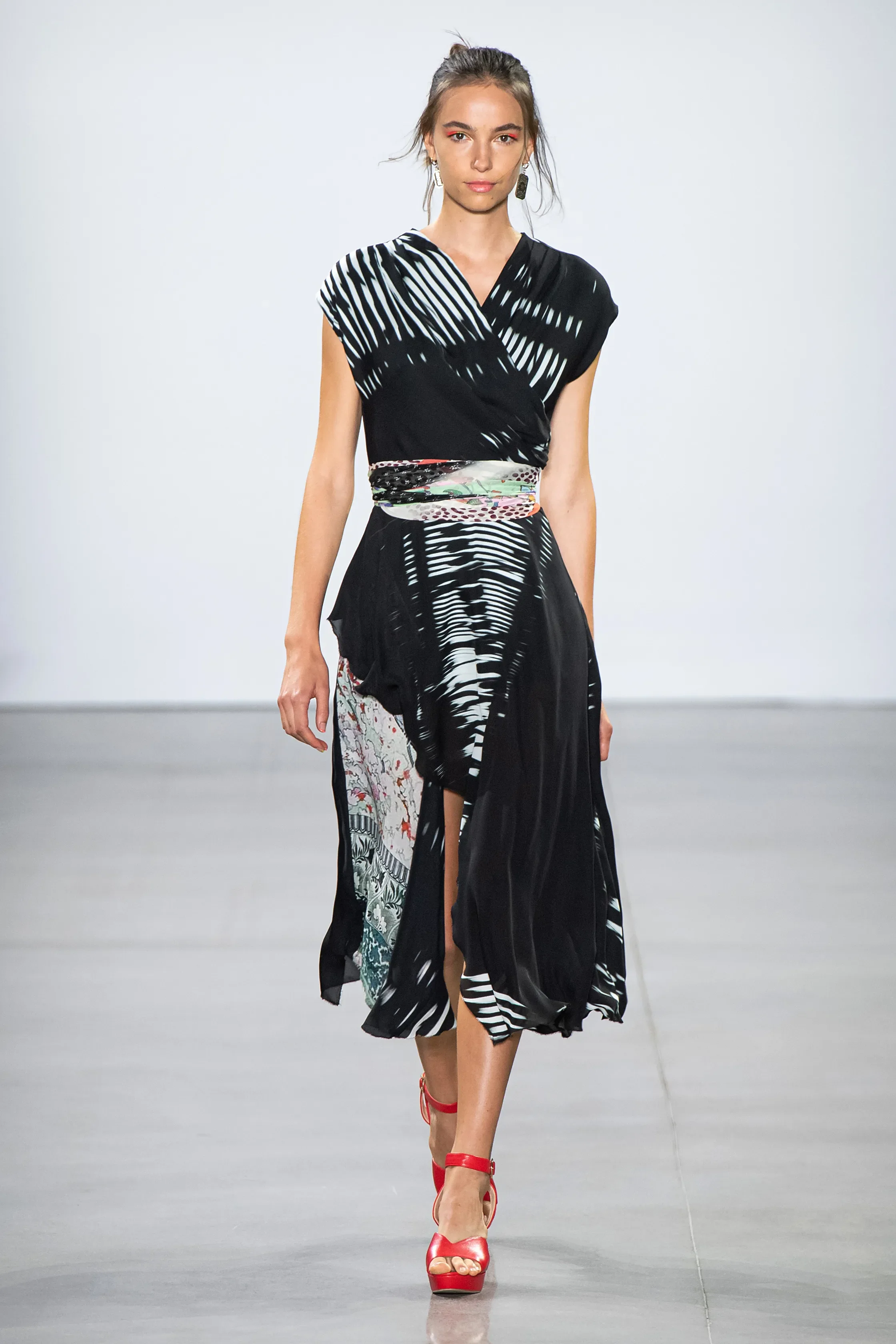 A female model walking on a runway wearing a black and white patterned dress with a high slit and red high-heeled sandals.