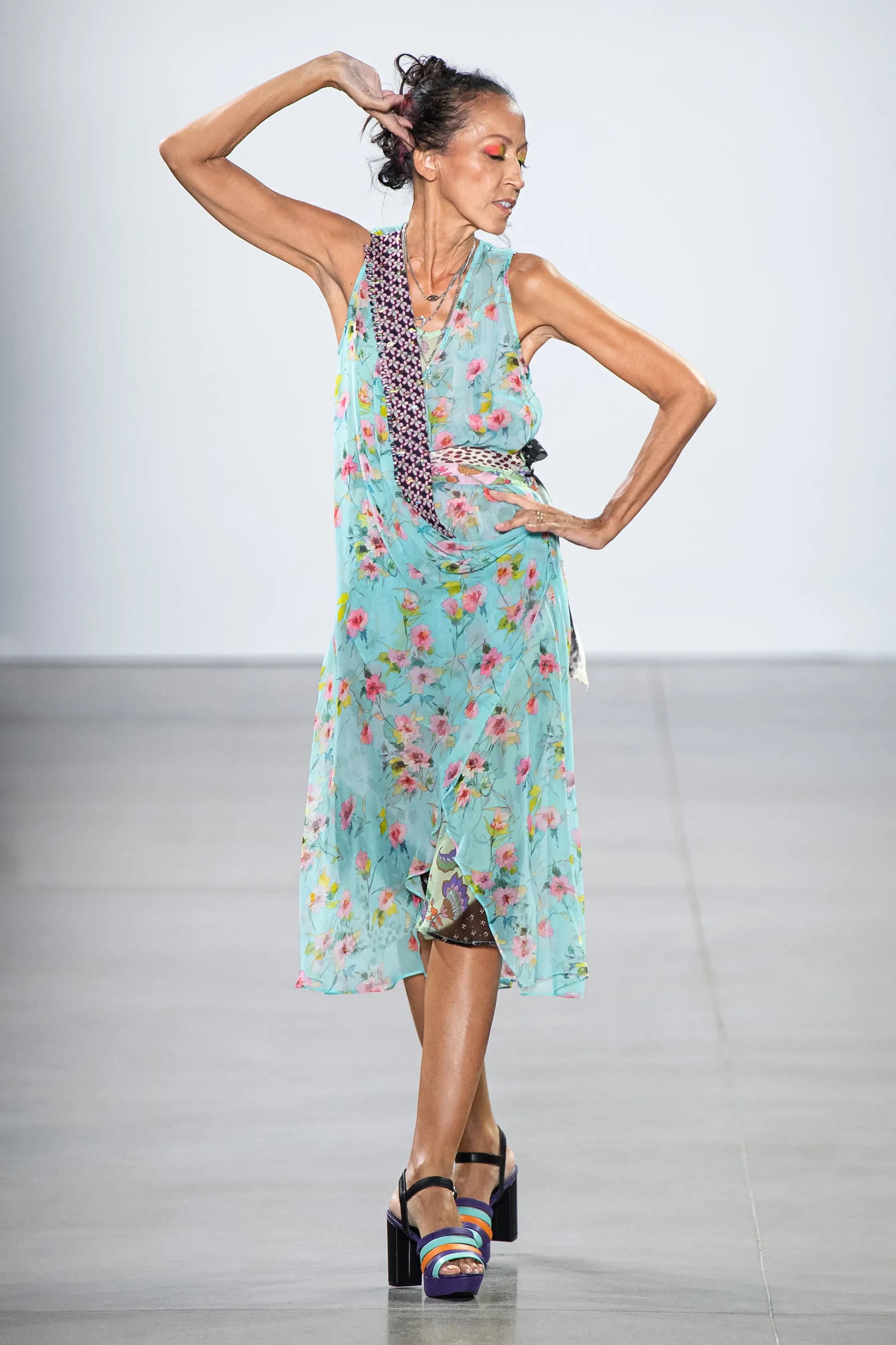 A woman in a colorful floral dress walking on a runway, wearing high-heeled sandals, with her hand on her hip and the other touching her head.