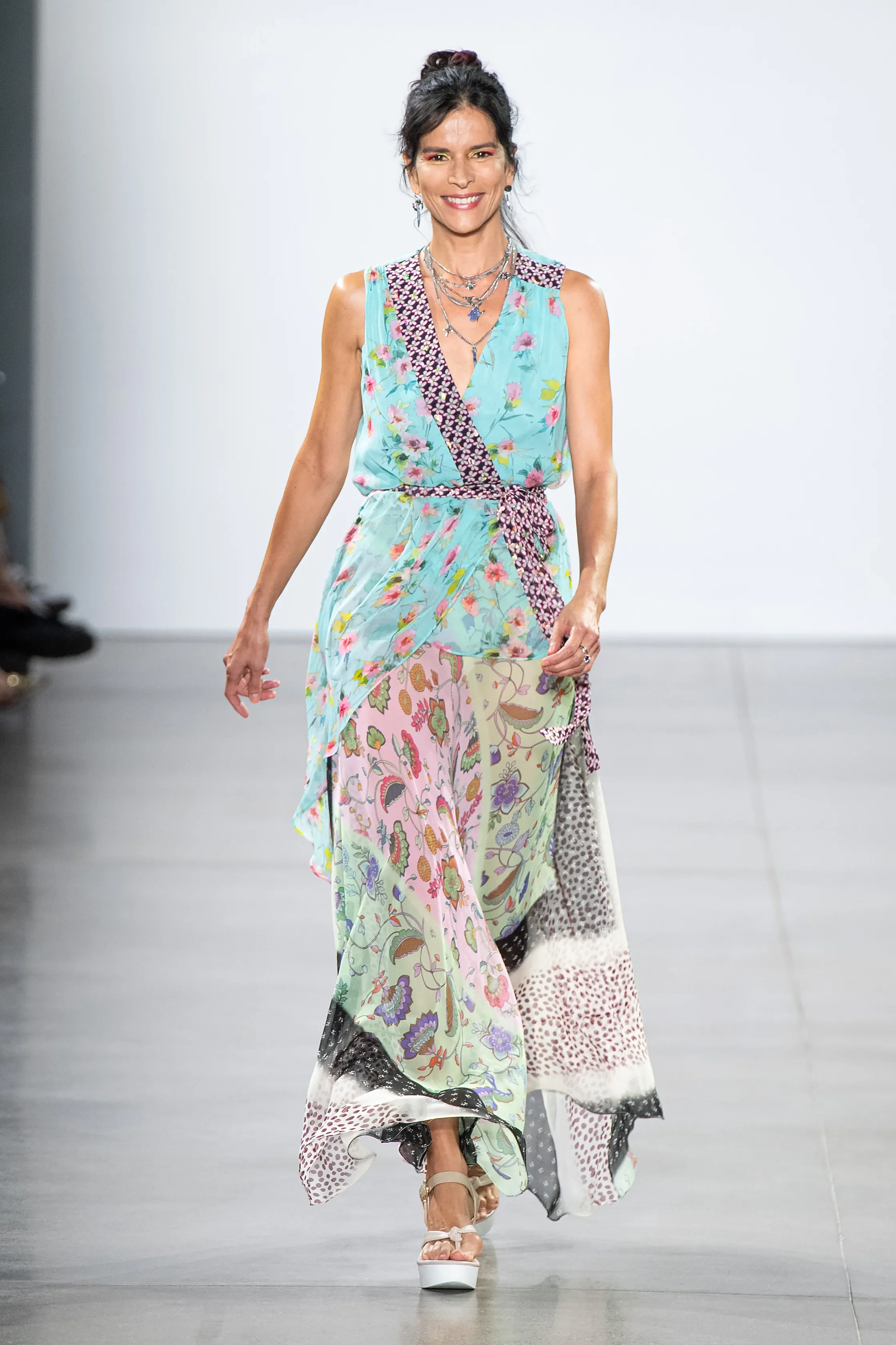 Woman walking on runway wearing a colorful, floral maxi dress with a mix of pastel and vibrant patterns, paired with platform heels and layered jewelry.