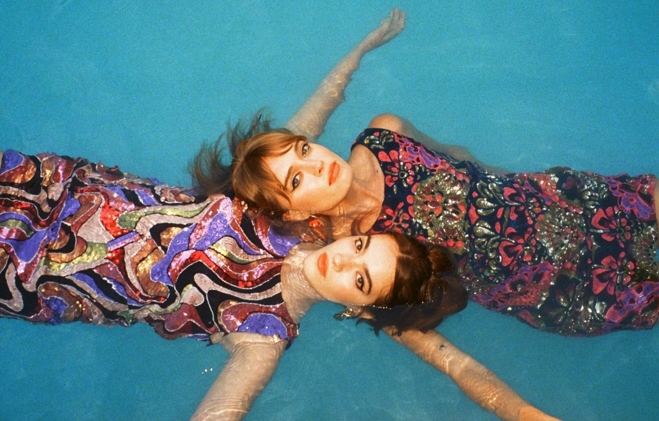 Two women in colorful, embellished dresses lying on a blue surface, with their heads close together, looking at the camera.
