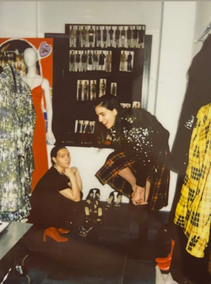 Two women in a clothing store, one sitting on a bench and the other bending over to adjust her shoe, with clothing and mannequins in the background.