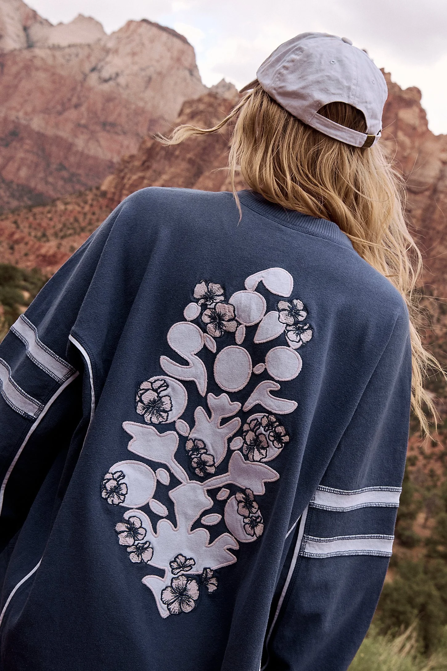 Back of a woman wearing a navy sweatshirt with floral embroidery, a white cap, and long blonde hair, in front of red rock formations.