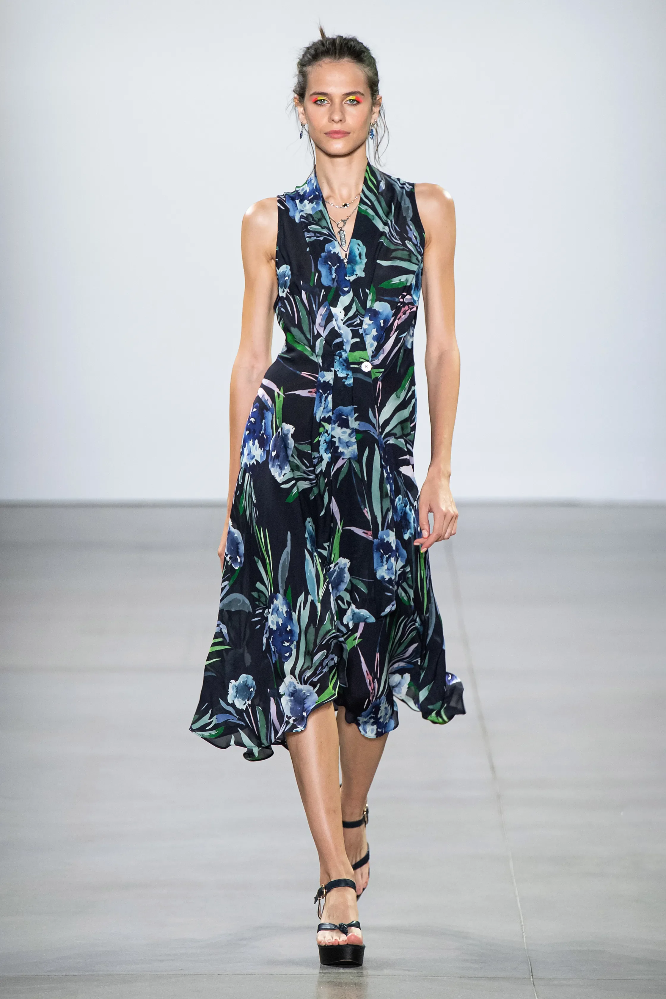 A female model walking on a runway wearing a sleeveless, flowy, navy blue dress with a colorful floral pattern, paired with black high heels.