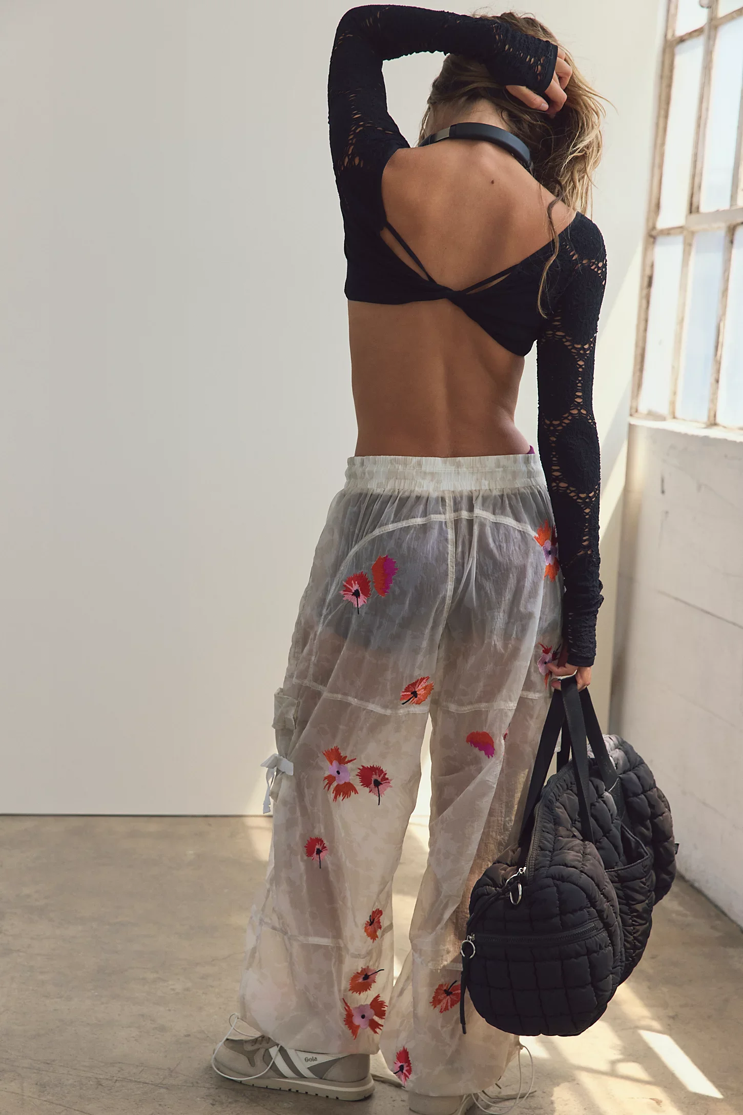 A woman standing indoors near a window, wearing a black lace long-sleeve top, sheer white pants with colorful floral embroidery, white sneakers, and holding a black quilted bag.