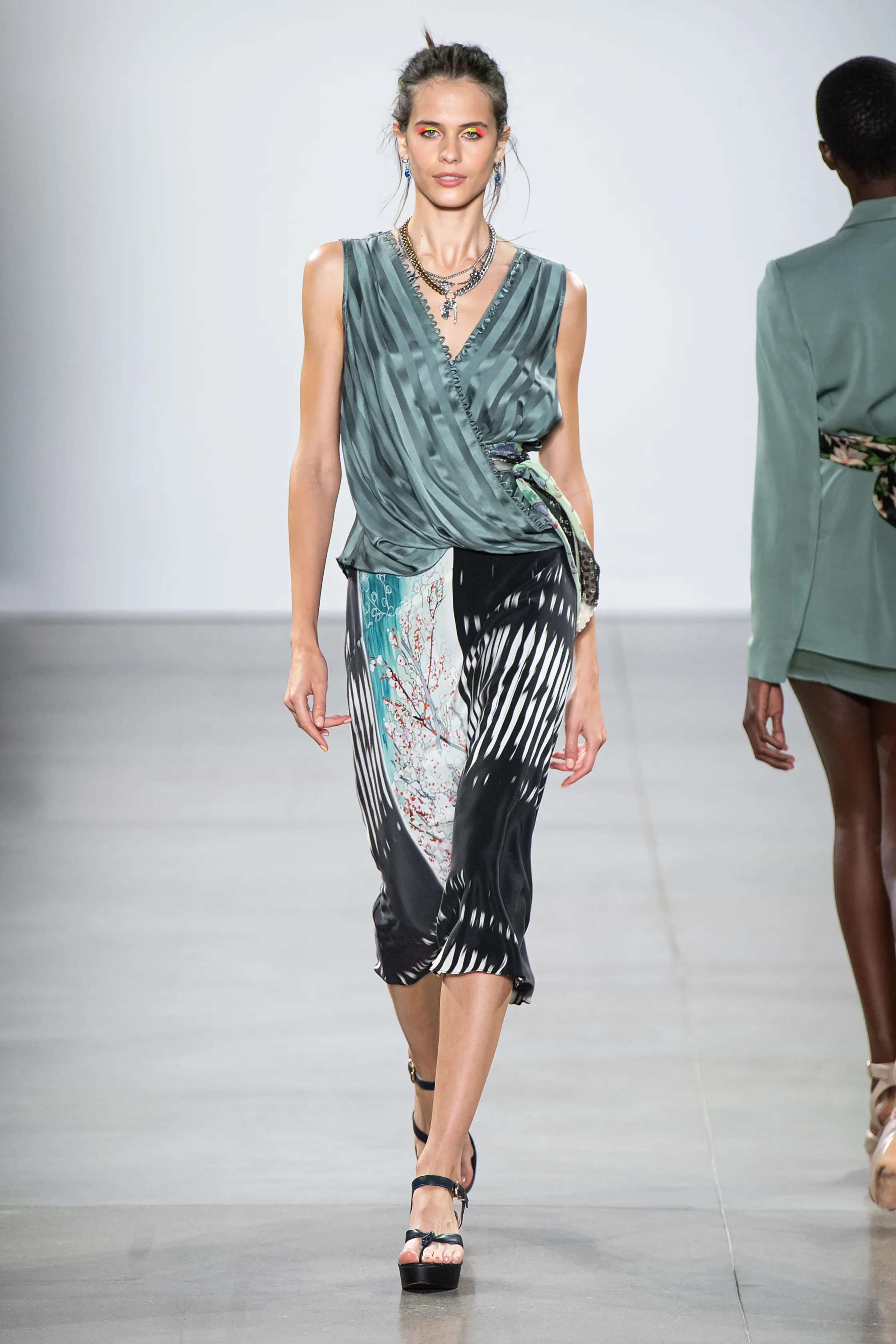 Model walking down runway in trendy, patterned outfit with platform heels, accessorized with layered necklaces and earrings.