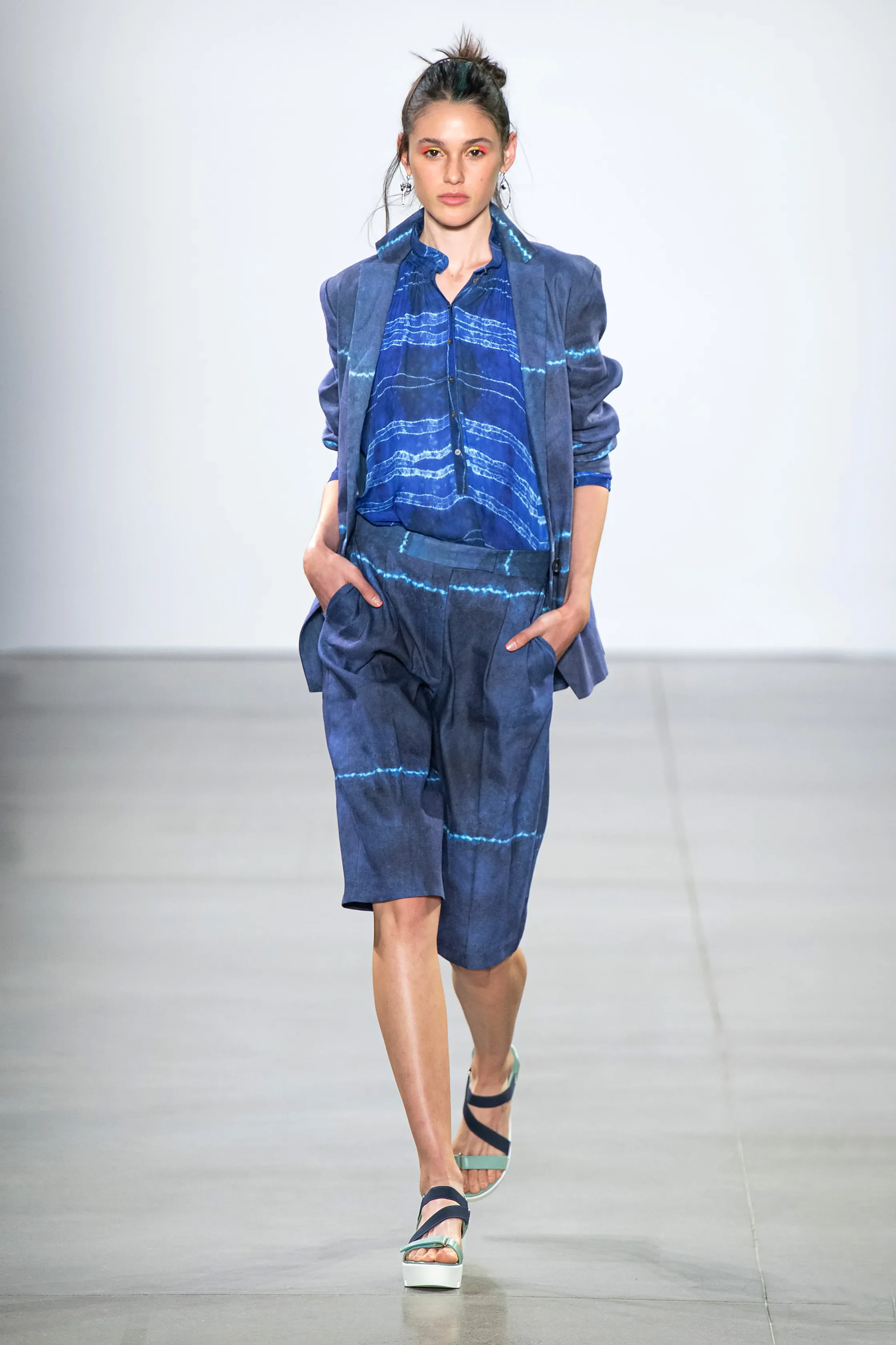 Female model walking on runway wearing a blue denim suit with a matching shirt and blazer, and platform sandals.