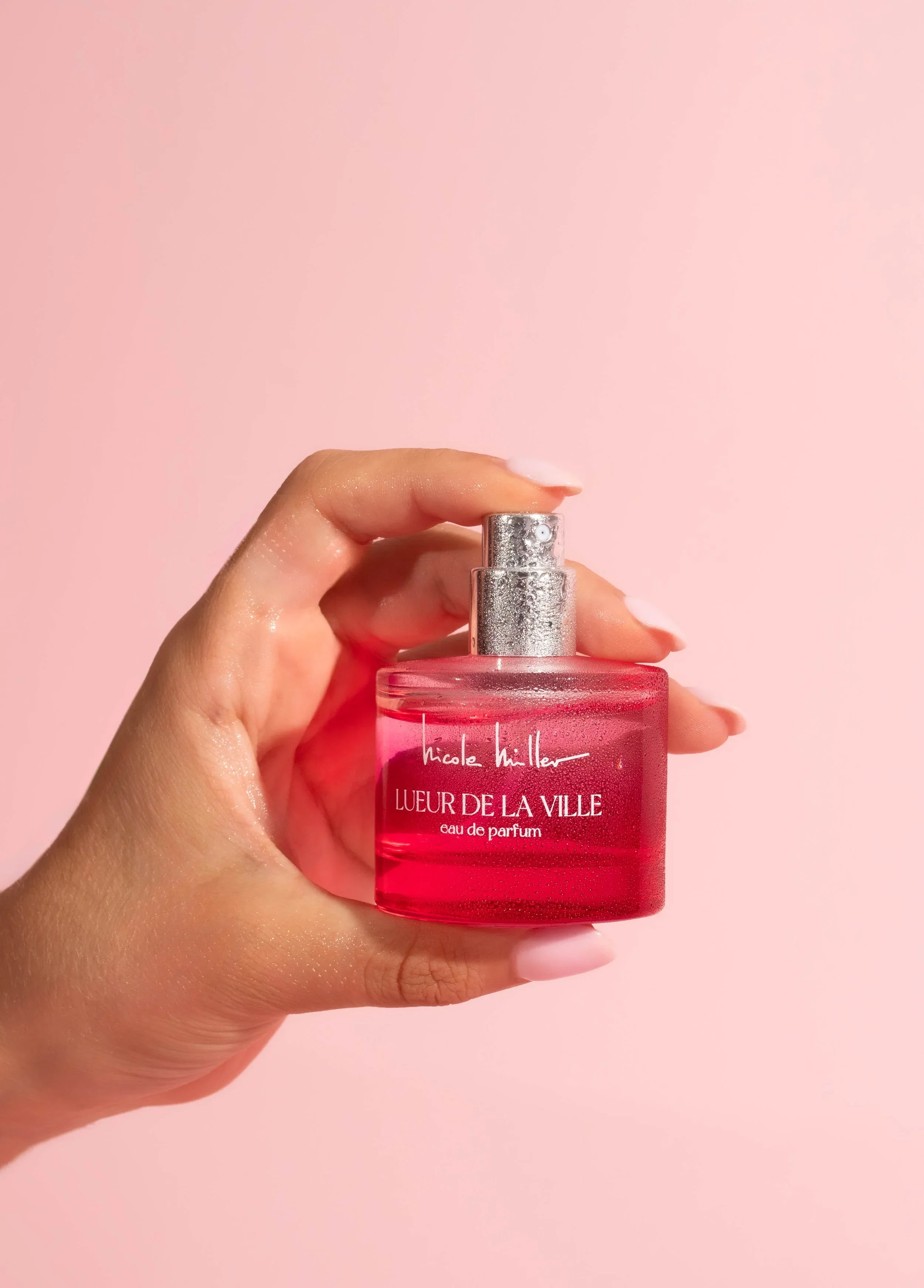 A hand holding a pink perfume bottle labeled 'Nicole Miller Lueur De La Ville Eau De Parfum', with a pink background.