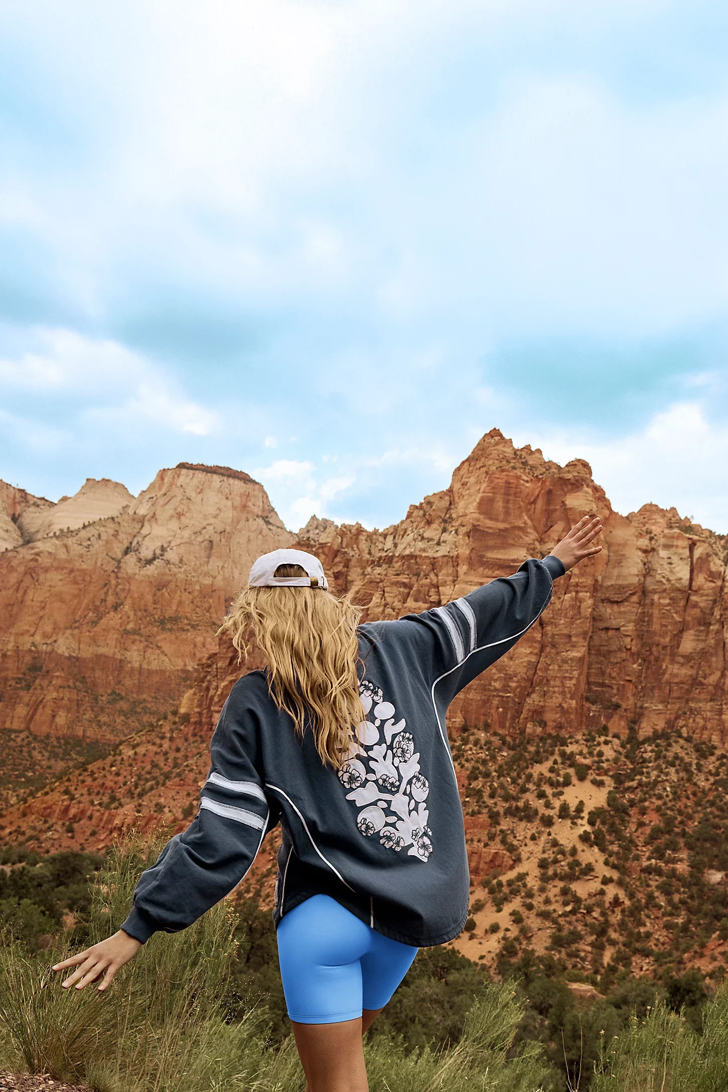 A woman with long blonde hair in a baseball cap and athletic jacket standing in front of red rock formations, posing with arms outstretched during the daytime.