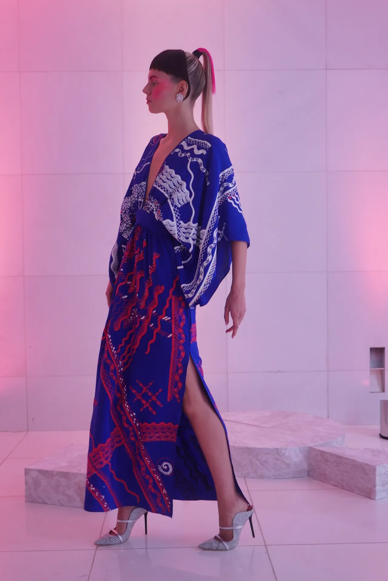A woman in a vibrant blue and red patterned dress with a high slit, stiletto heels, and a styled hairdo stands against a pink-lit tiled wall.