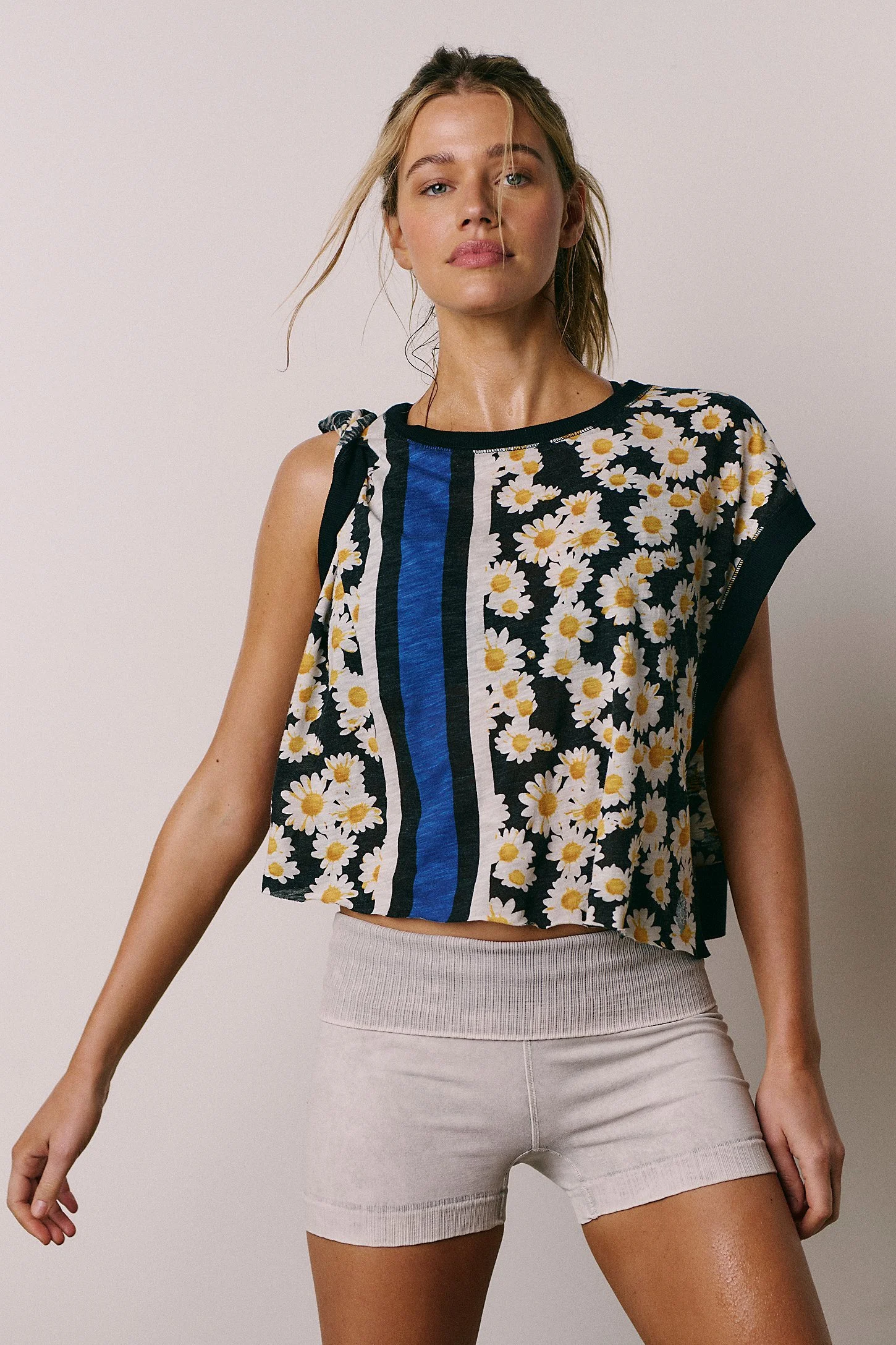 A woman in a black, white, blue, and yellow floral sleeveless top, paired with beige shorts, standing against a plain background.