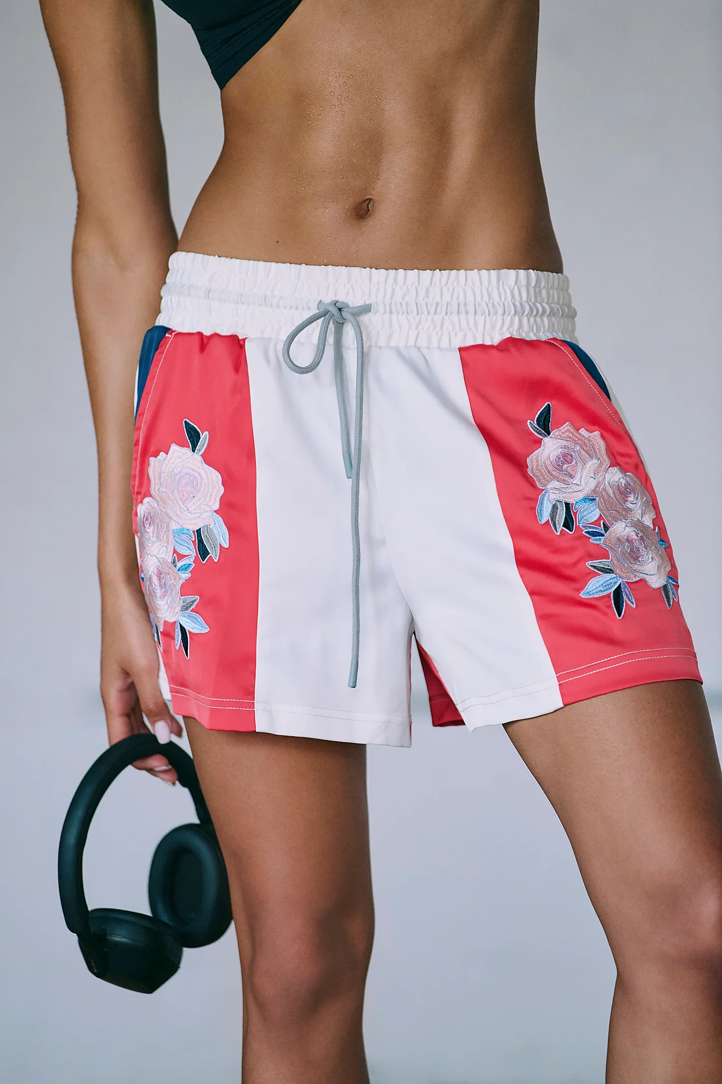 Close-up of a woman's lower torso wearing swim shorts with floral embroidery and holding a pair of black headphones