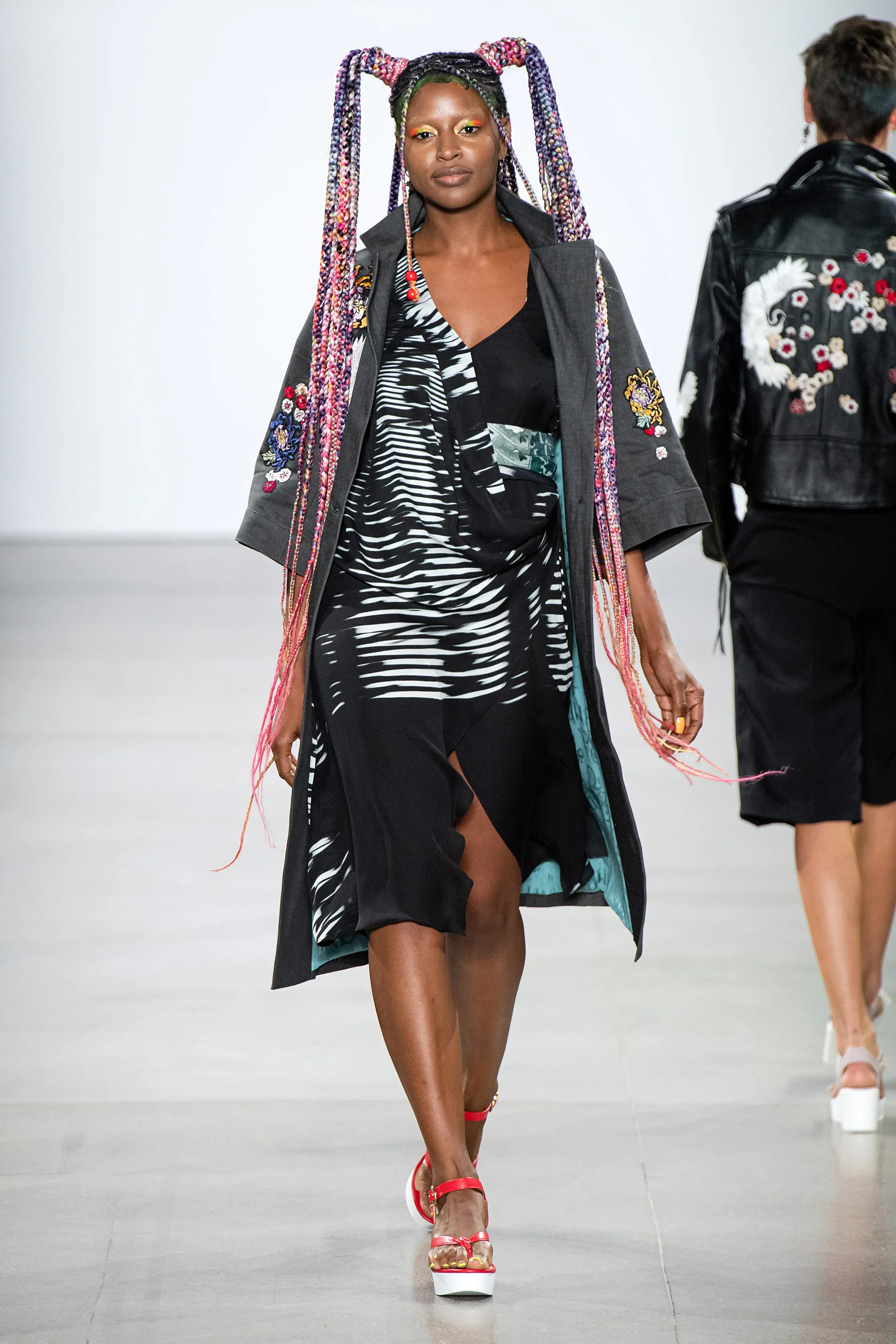 A woman walking on a runway during a fashion show wearing a black and white patterned dress with a zipper slit, open knee-length black coat with embroidered floral patches, red high-heeled sandals, and colorful braided hair styled in long pigtails.