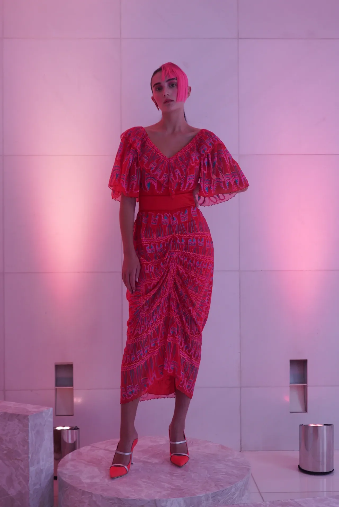 A mannequin dressed in a vibrant pink and red patterned dress with ruffled sleeves, standing on a marble platform against a pink-lit wall.