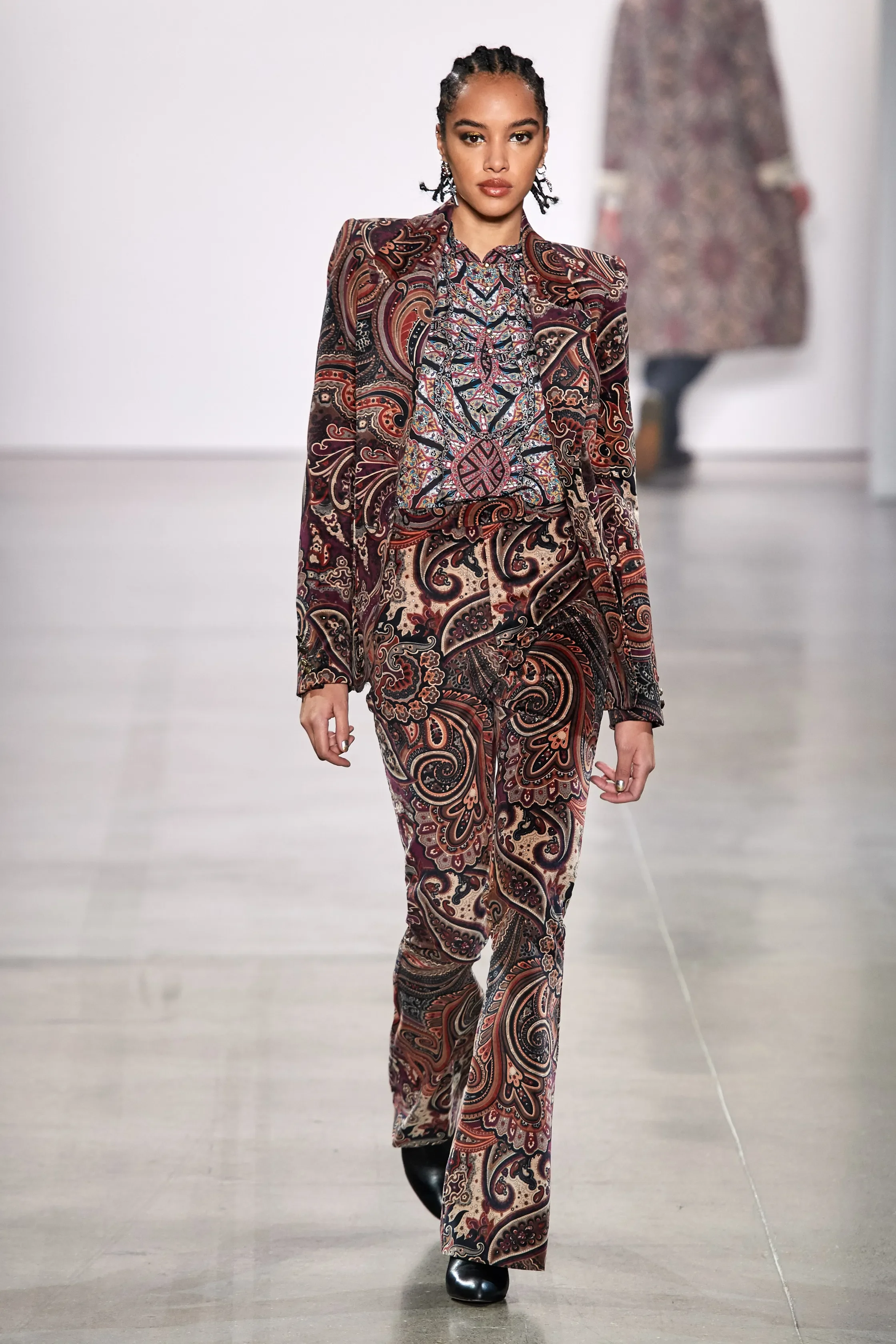Model walking on runway wearing a patterned suit with ornate paisley and floral designs in shades of brown, red, black, and cream.